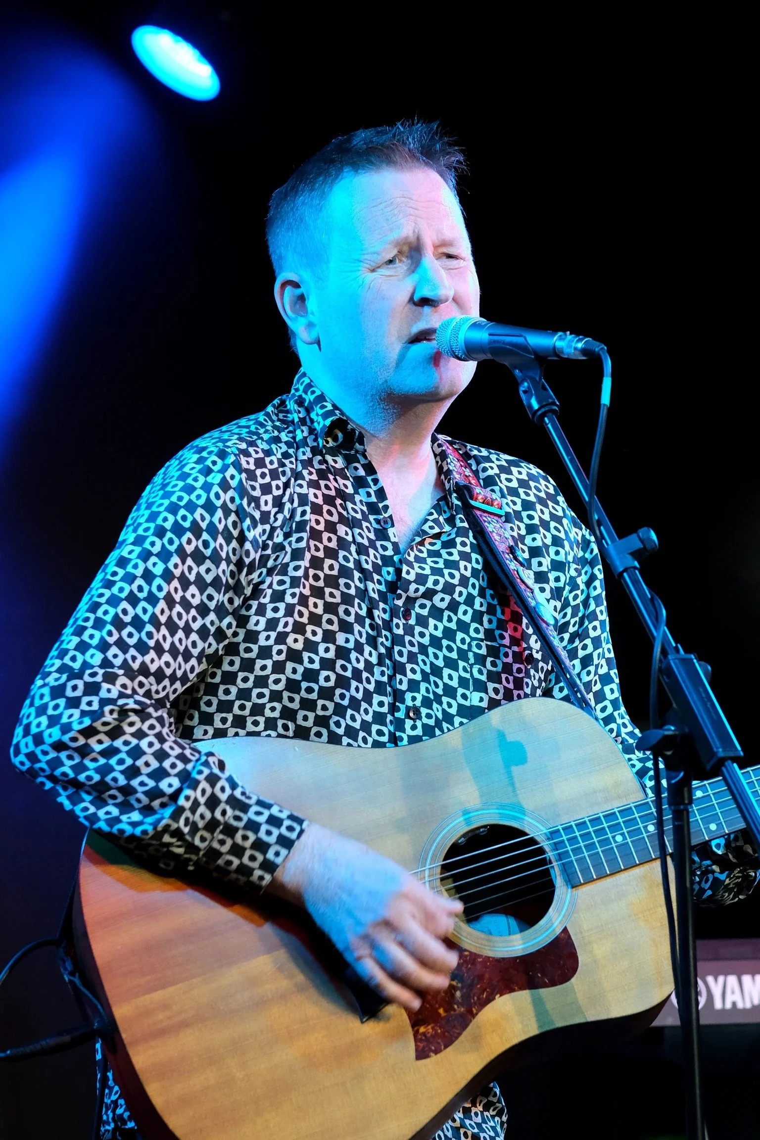 A man singing into a microphone while playing an acoustic guitar on stage, illuminated by blue stage lighting.