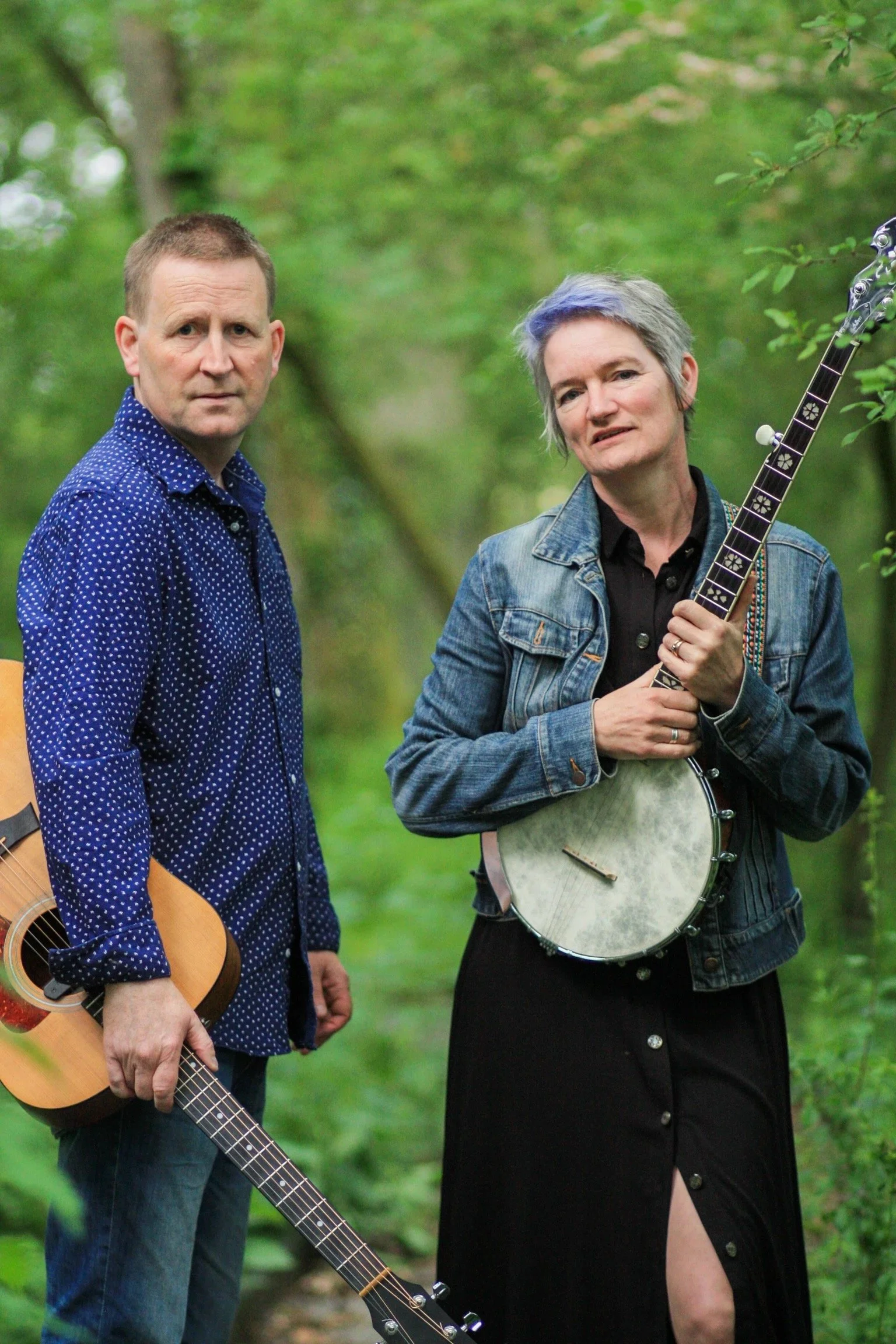 Two people standing outdoors in a green wooded area, each holding a musical instrument. The person on the left holds an acoustic guitar, and the person on the right holds a banjo. The woman on the right has short gray hair with a hint of blue, and is