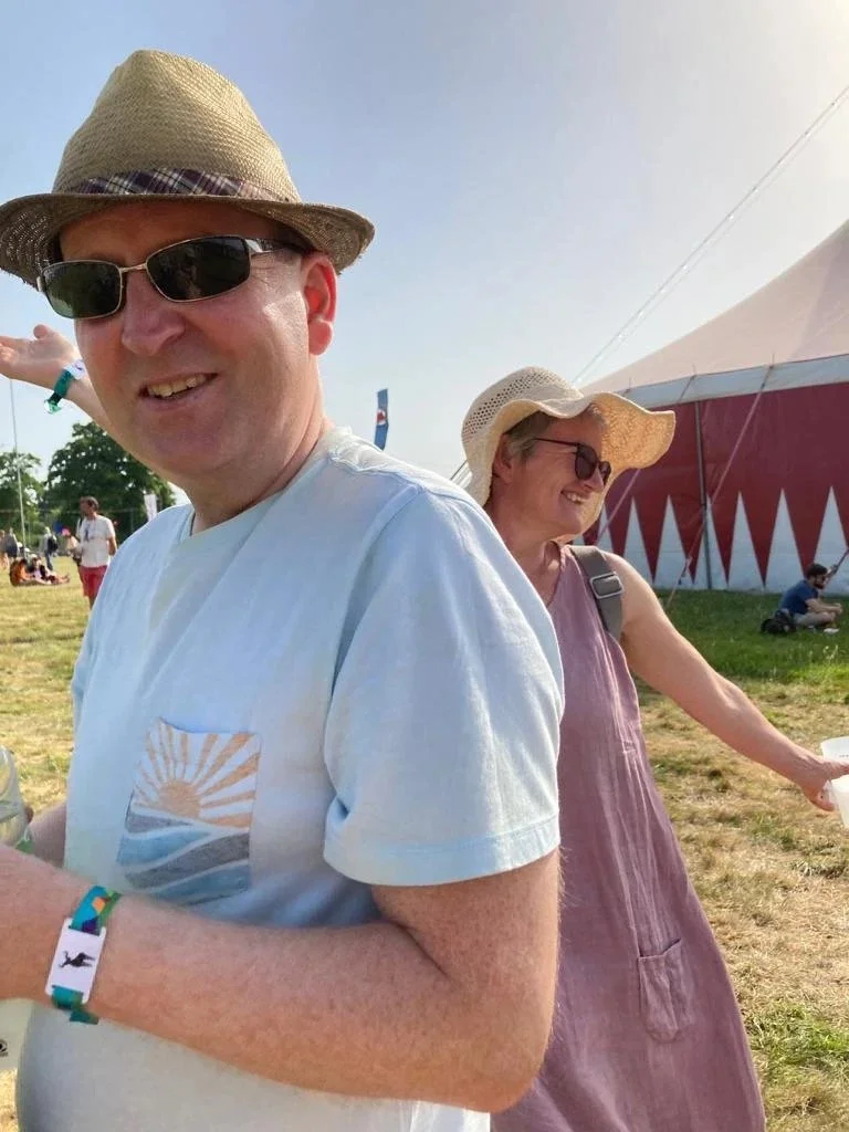 Two people outdoors at a festival or fair, wearing hats and sunglasses, with a large tent in the background.