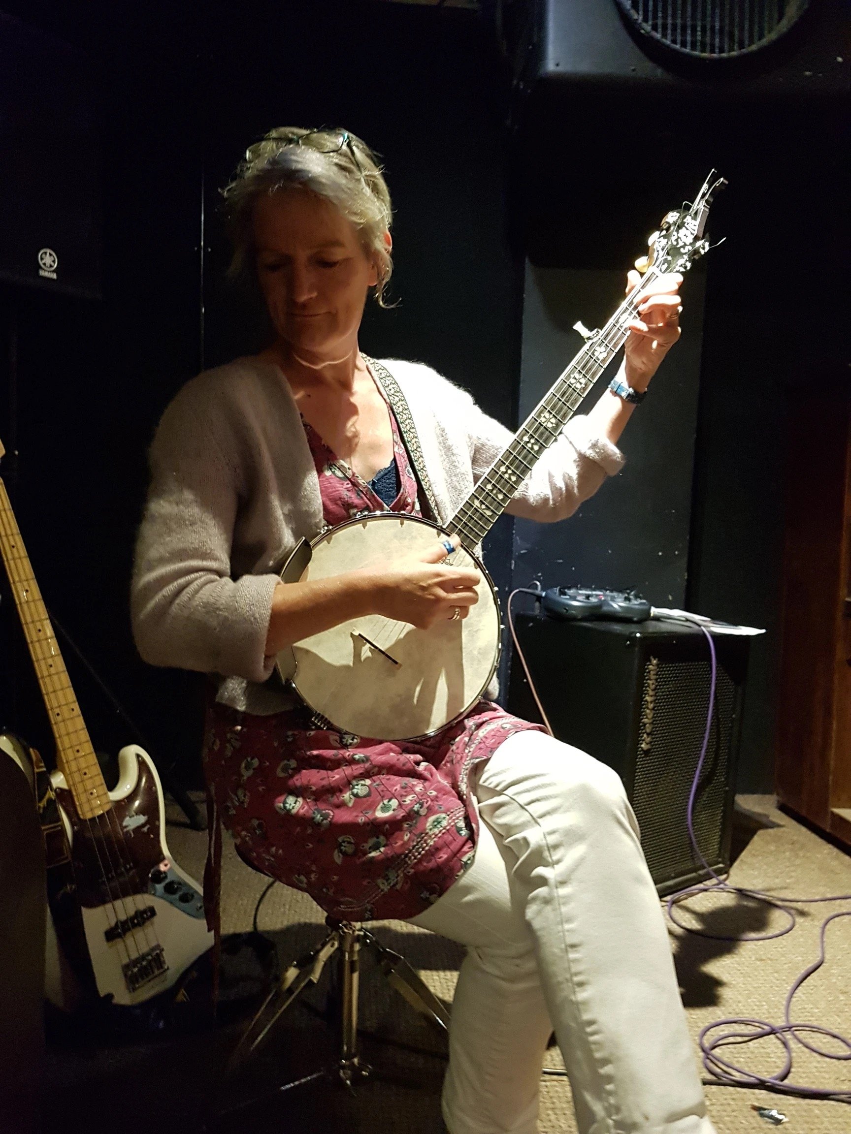 A woman playing a banjo, seated on a drum throne in a dimly lit room with musical equipment around her.