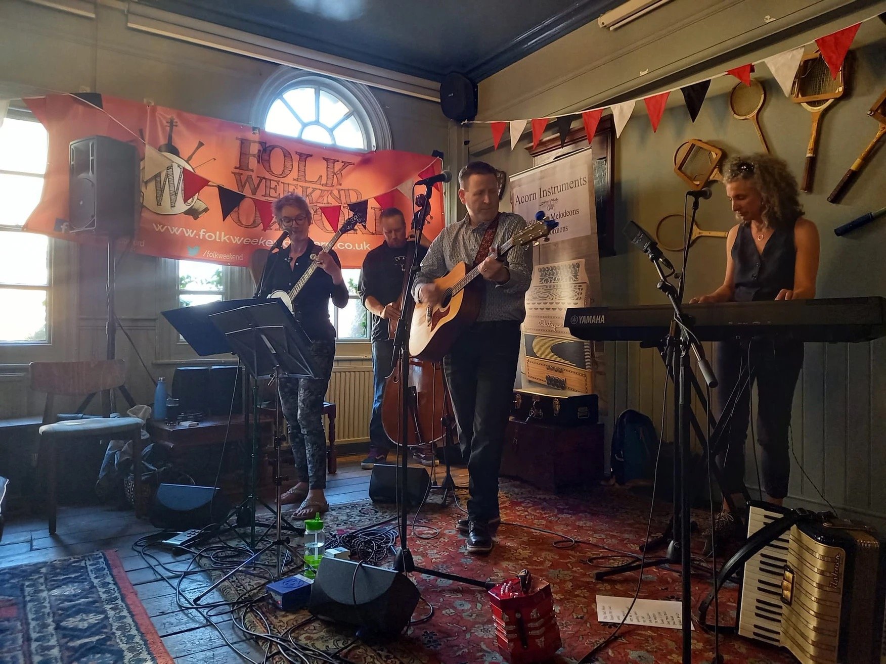 A four-piece folk band performing on a stage with acoustic and electric instruments, including guitars, keyboard, and ukulele, at an indoor event with musical banners and hanging decorations.