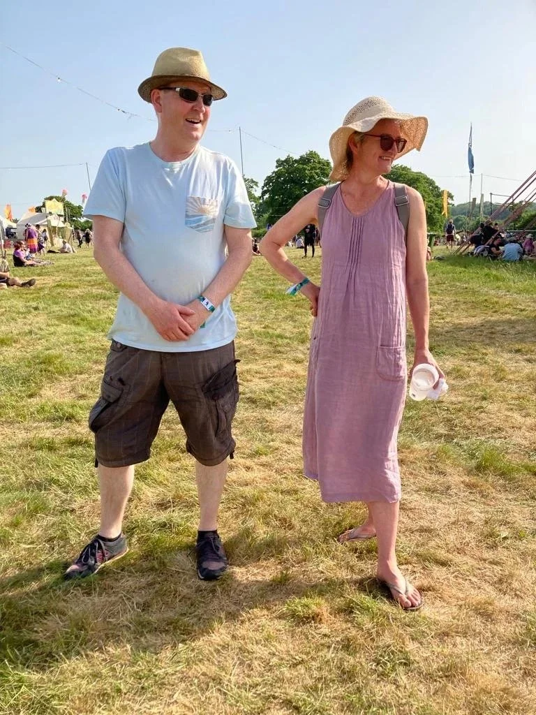 A man and woman standing outdoors at a festival on a sunny day, wearing hats and sunglasses, with festival tents and people in the background.