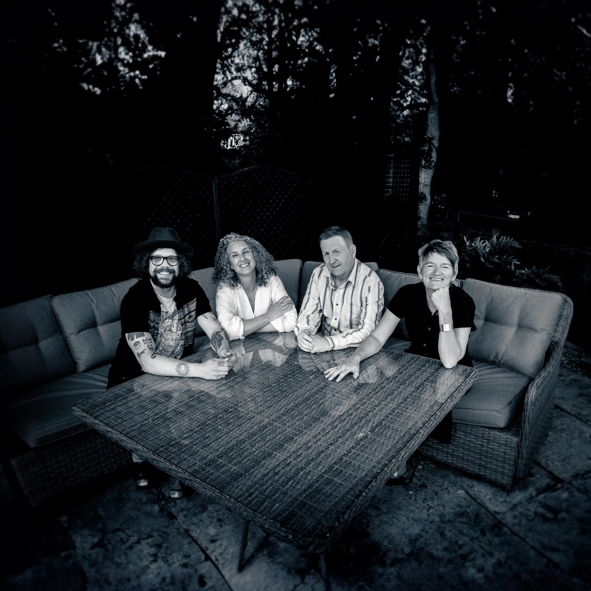 A black and white photo of four people sitting at an outdoor table on a patio. They are smiling and looking at the camera. Behind them are trees and a fence.