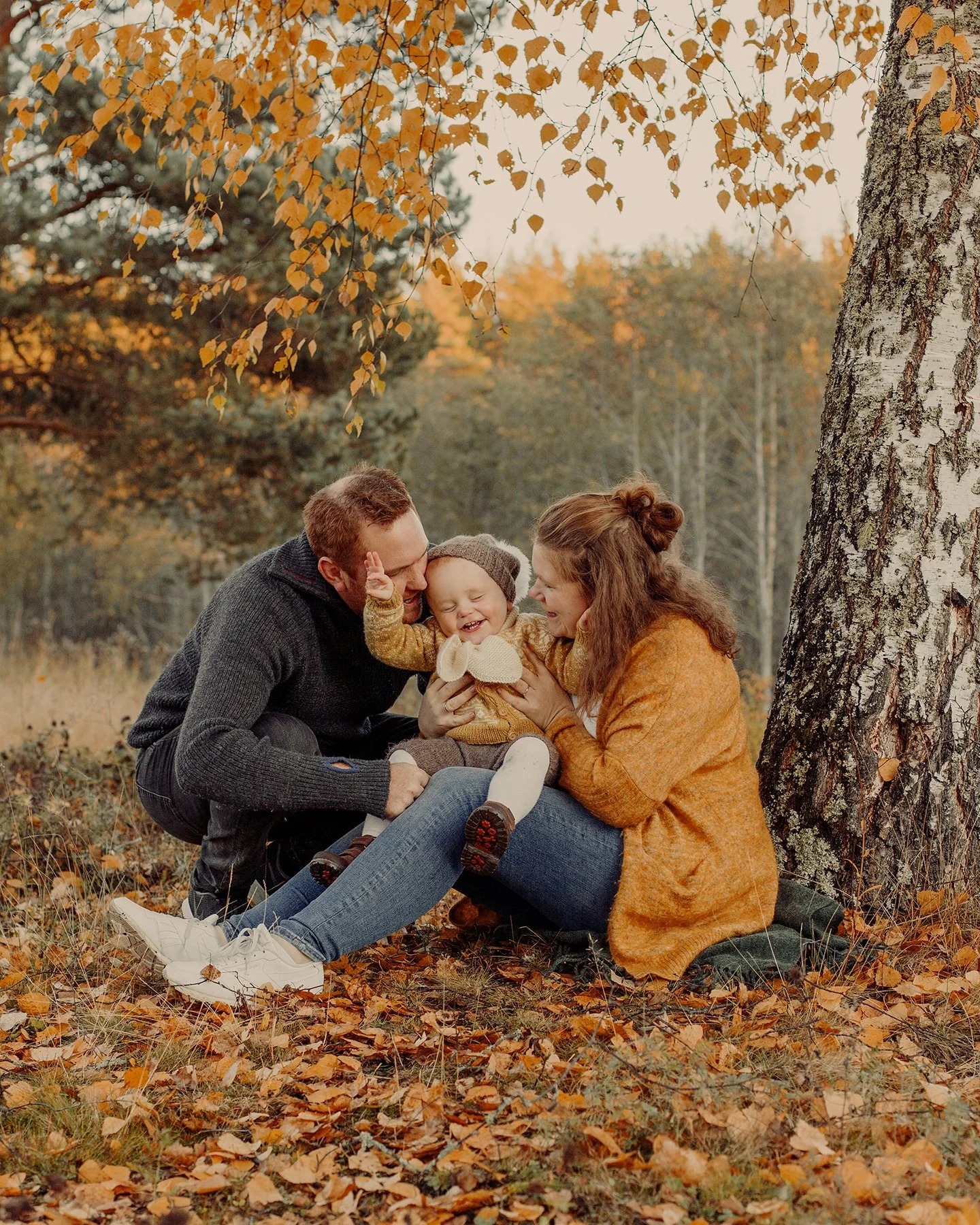 Barnefotografering om høsten. Mor og far tøyser med barnet som sitter på fanget under et tre.