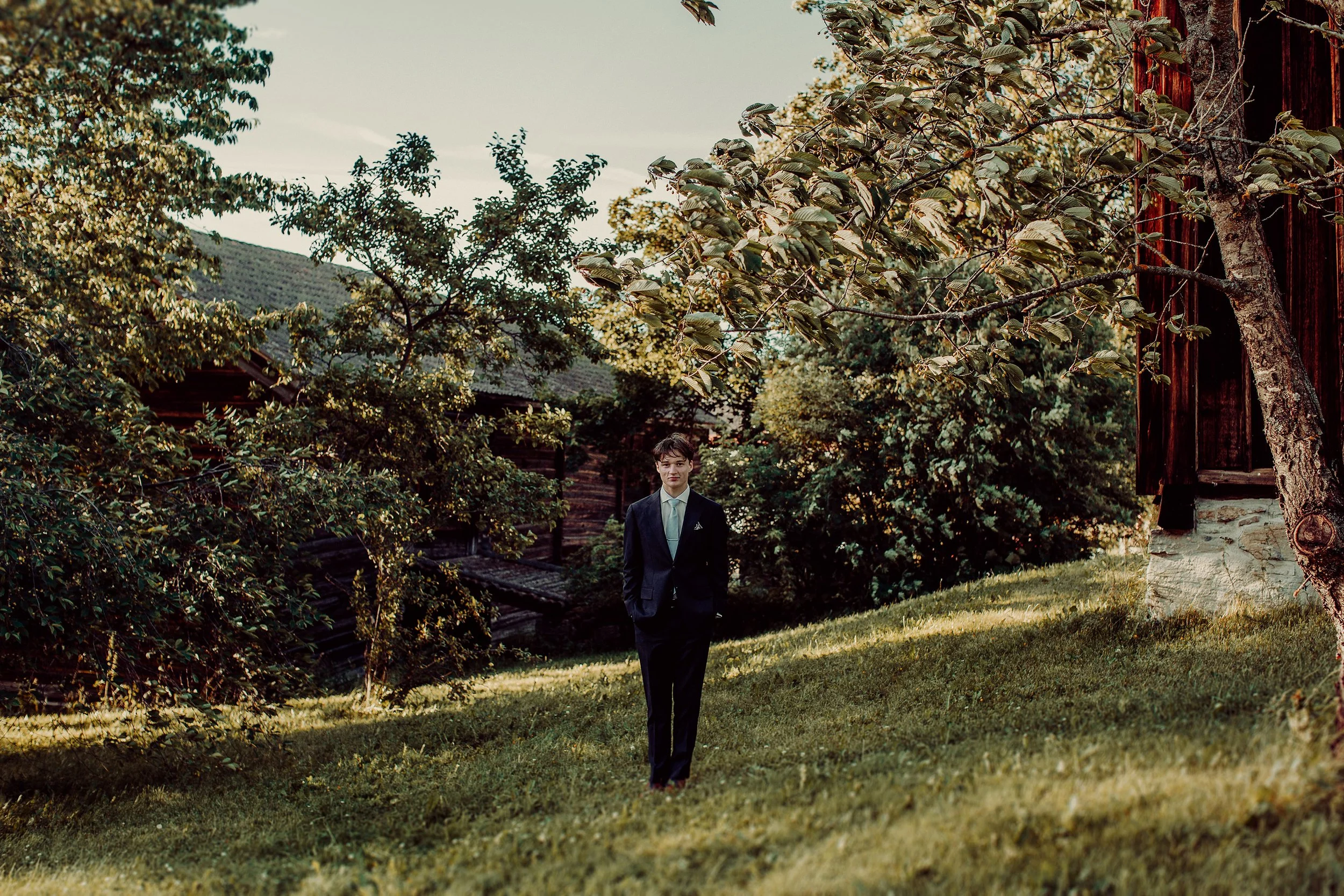 Konfirmantfotografering av gutt I dress omkranset av gamle trær og bygninger