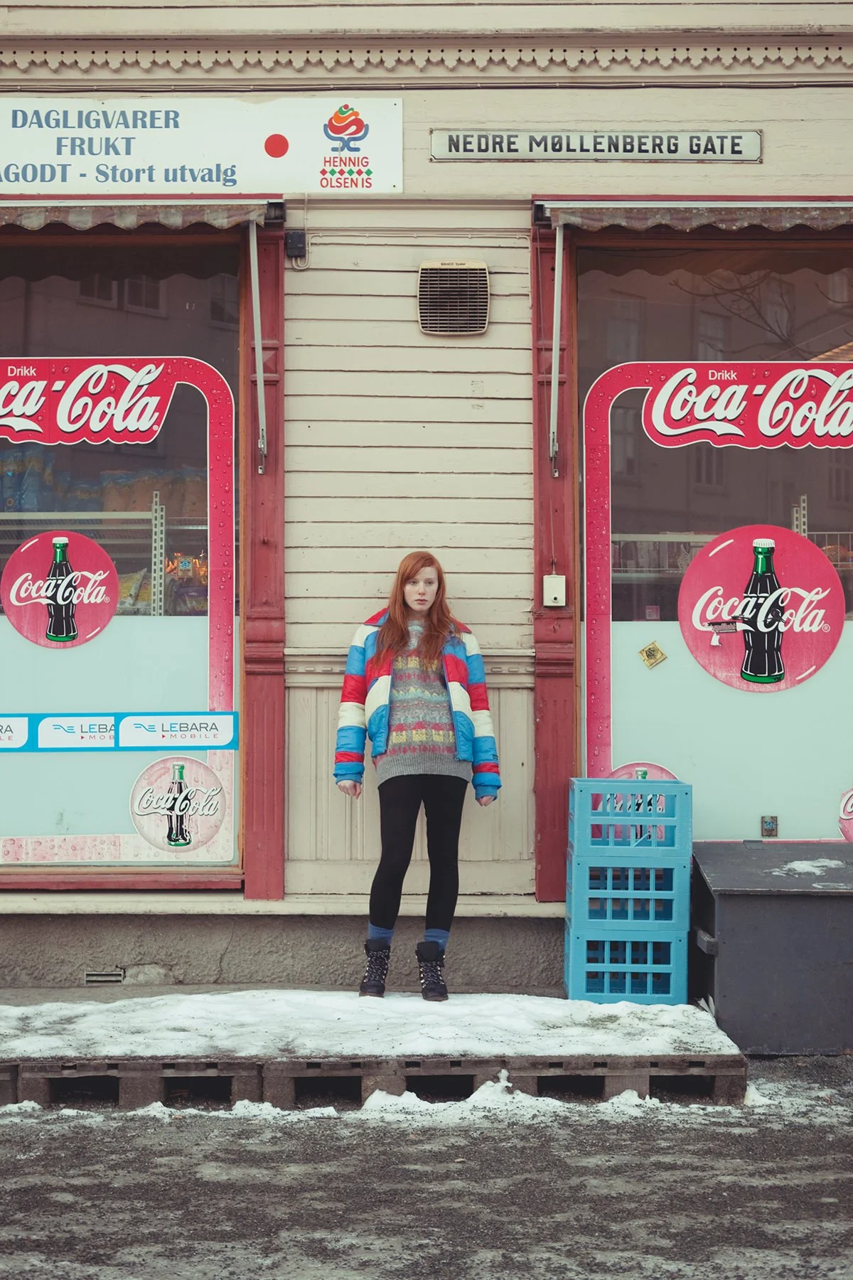 Ung jente med rødt hår og boblejakke står utenfor en sliten butikk på en kald vinterdag.