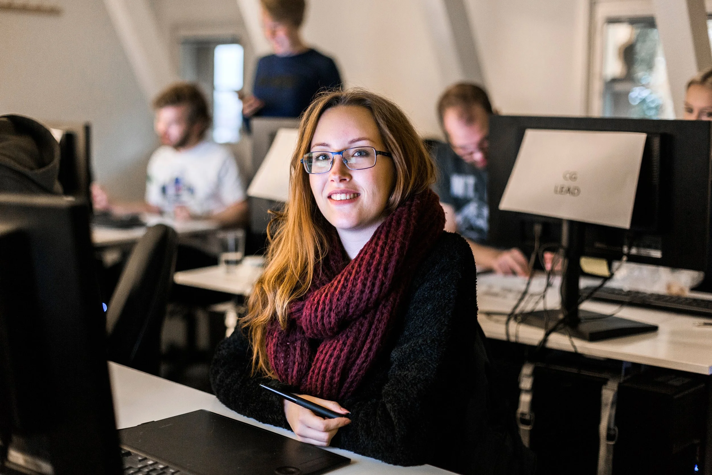 Kvinnelig student sitter ved datamaskinen og smiler til kamera.