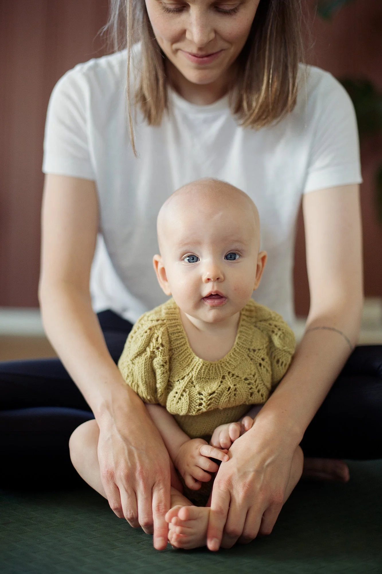 Mor og baby gjør yoga. Babyen sitter på gulvet og moren holder hendene sine rundt barnet.