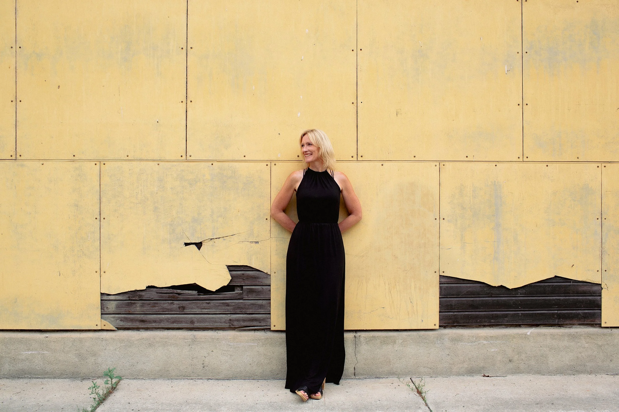 Kvinnelig leder ikledd kjole står avslappet inntil en gul metallvegg. To av platene er ødelagt.