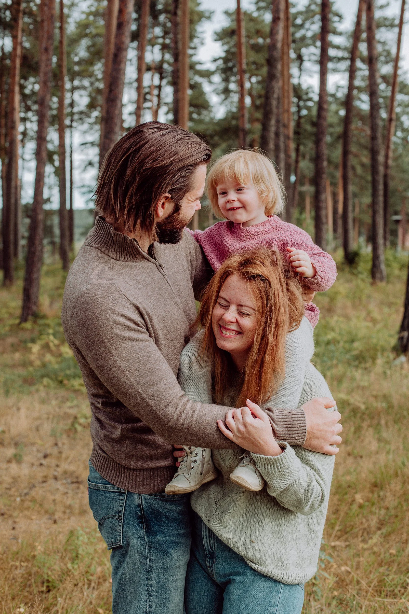 Et barn sitter på skuldrene til sin mor og tuller med far som holder rundt mor på familiefotografering ved Mjøsa.