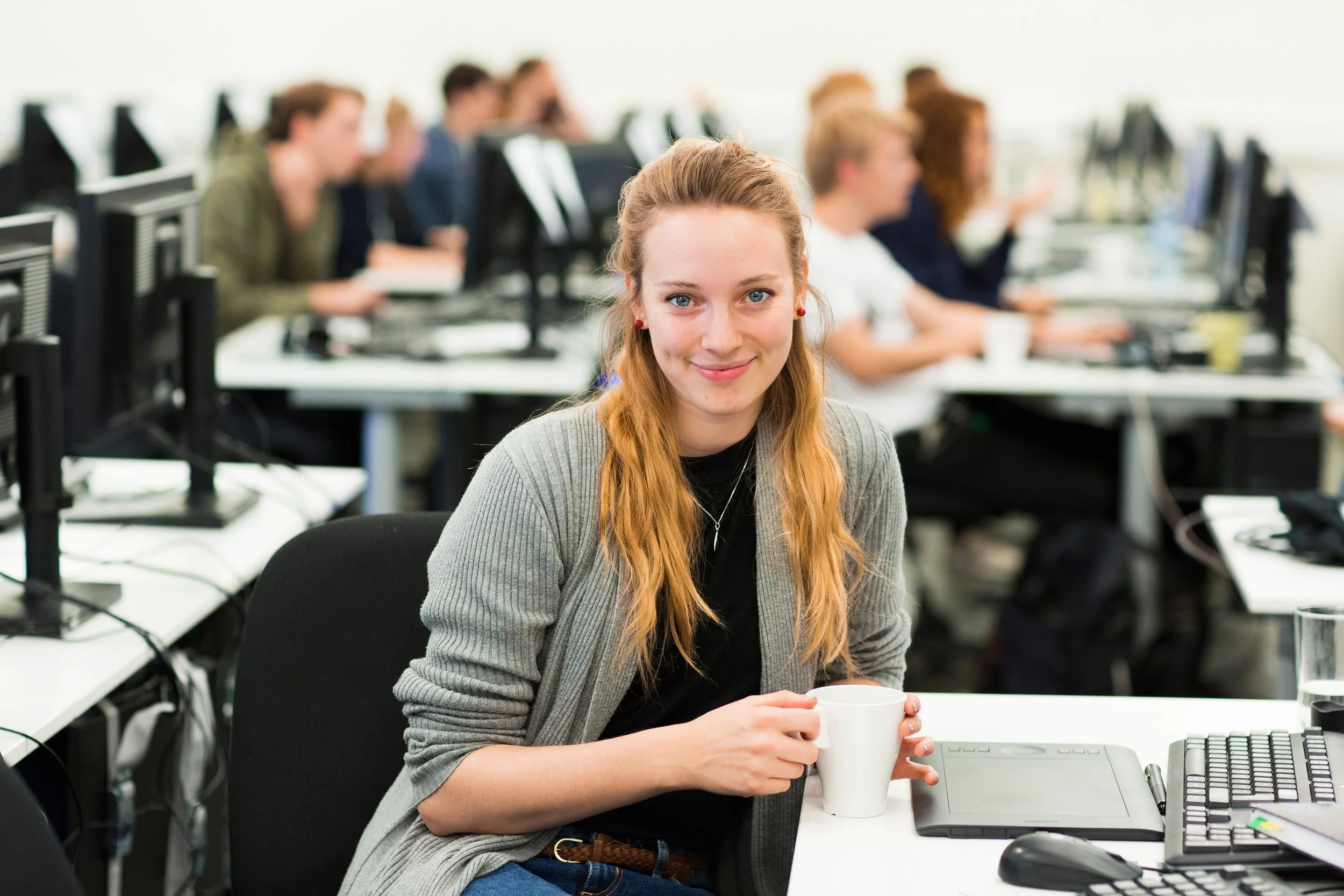 Portrettbilde i miljø av kvinnelig student som sitter ved pulten med en kaffekopp.