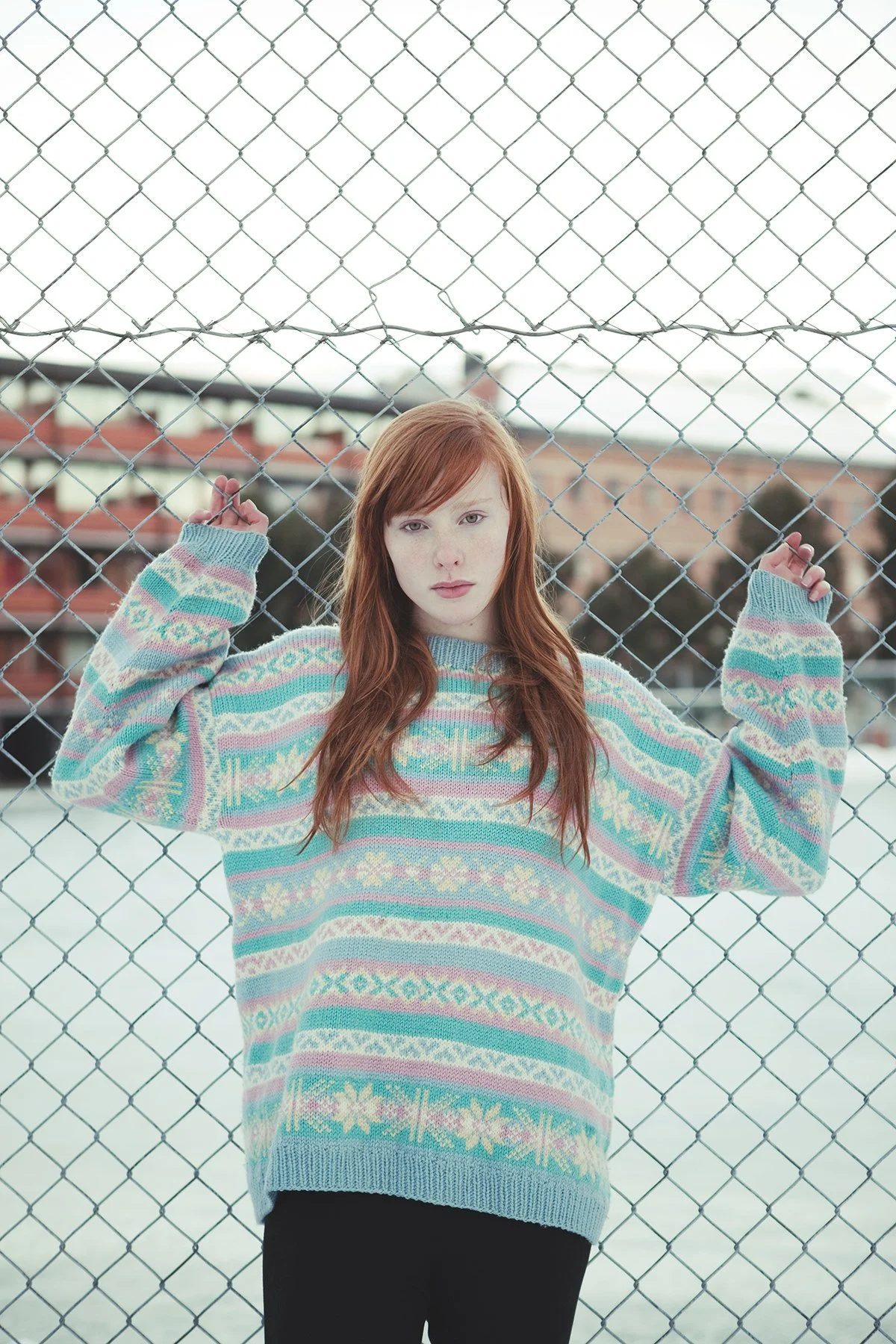 Ungdomsportrett av jente med rødt hår som lener seg inn til et nettinggjerde mens hun holder fast i gjerdet med hendene. Det er vinter og ser kaldt ut.