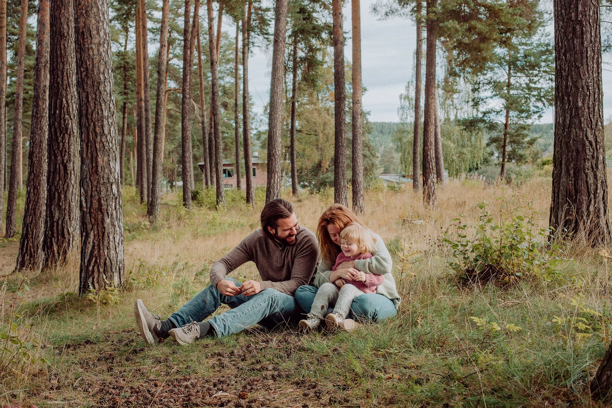 Familiefotografering utendørs i naturlig lys. Familie på tre, to voksne og ett barn, sitter på bakken sammen blant furutrær.