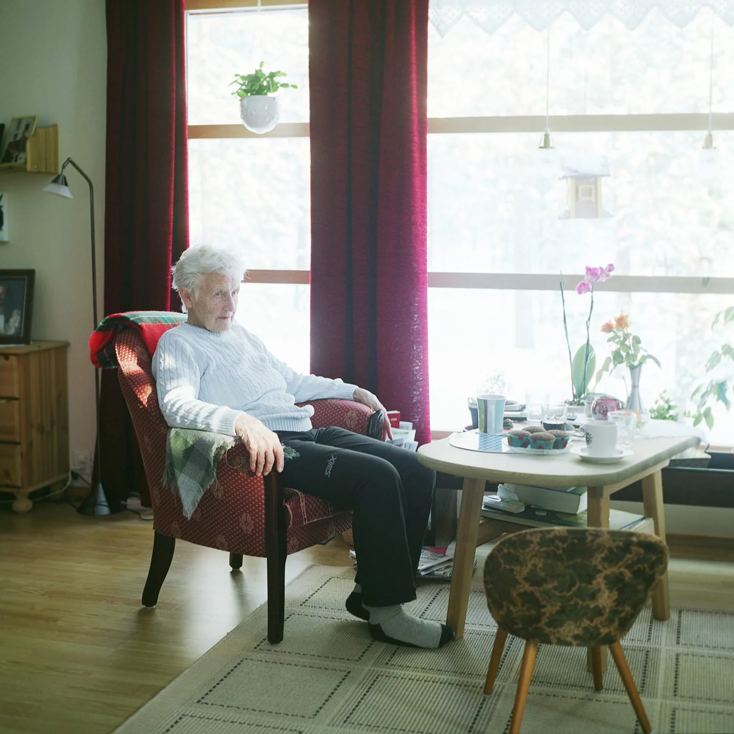 Eldre kvinne sitter i en lenestol foran et stort vindu med lysinnslipp.