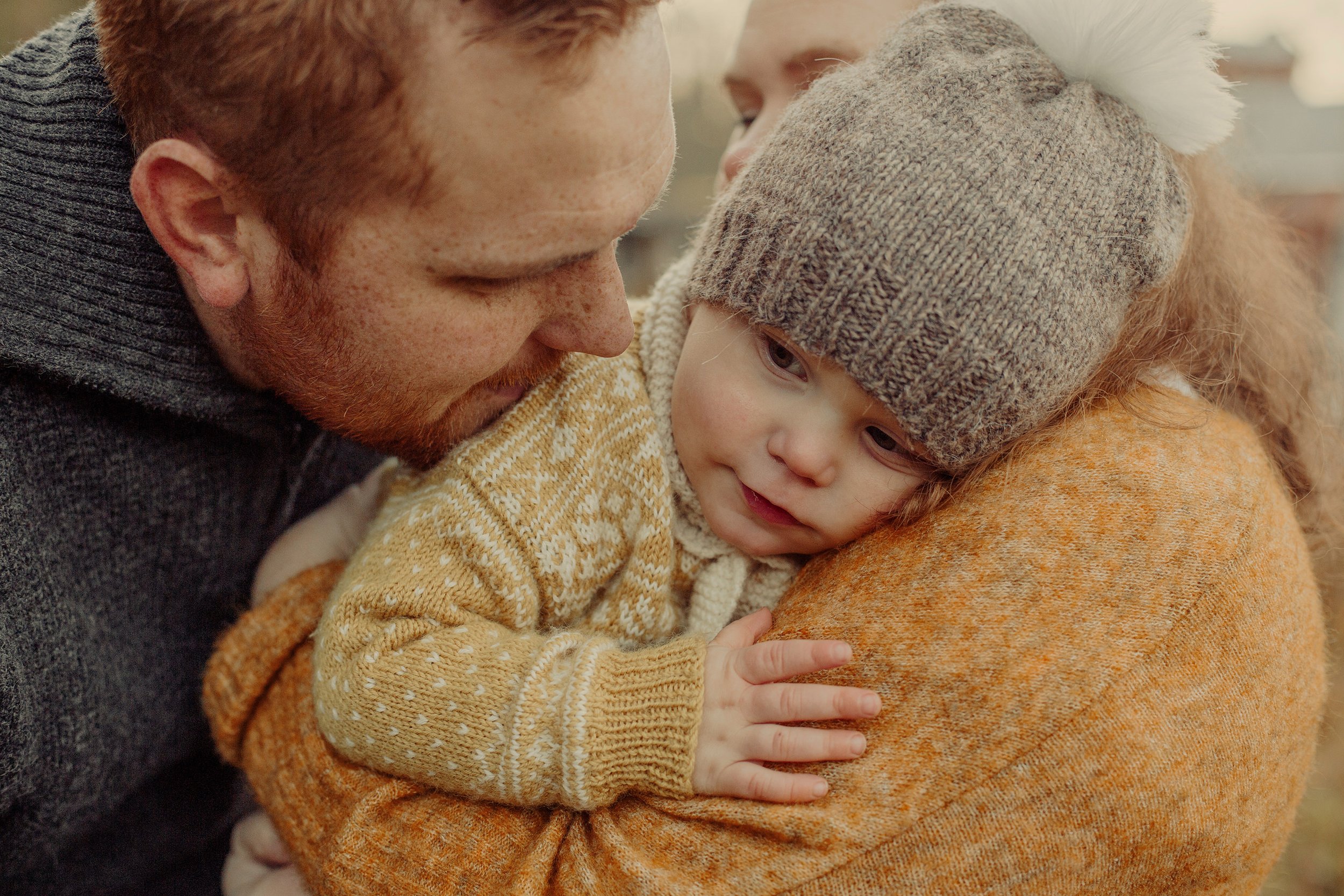 Et lite barn hviler på skulderen til sin mor. Faren lener seg inn og gir barnet en klem.
