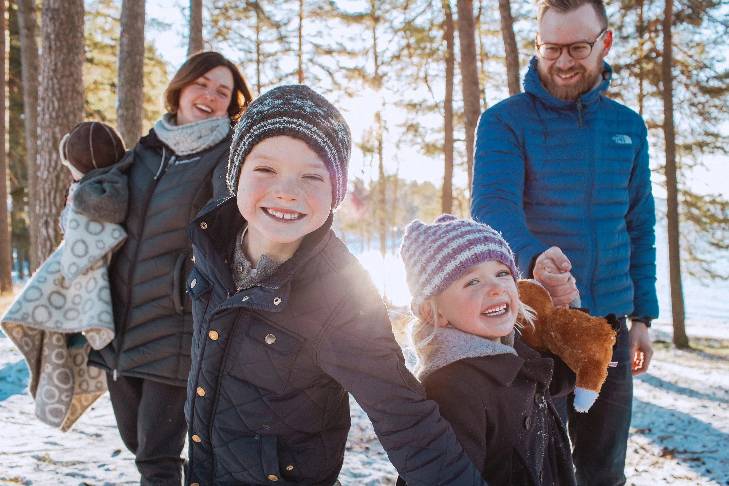Familiefotografering utendørs på vinteren. Familien holder hender og går på rekke mot kamera mens de smiler og ler.
