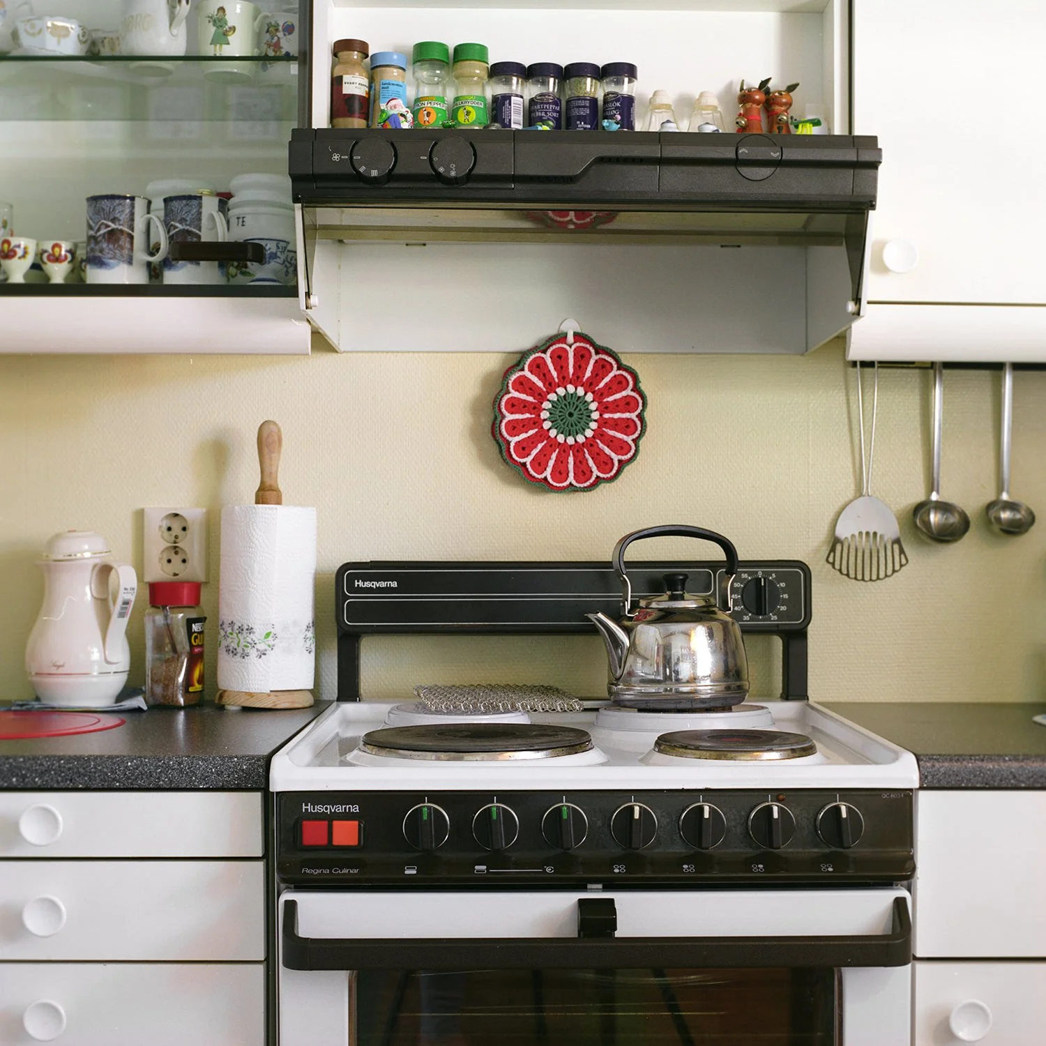 Utsnitt av interiør på kjøkken. Gammelt kjøkkeninteriør med en kaffekjele på kokeplate, og rød gryteklut på veggen.