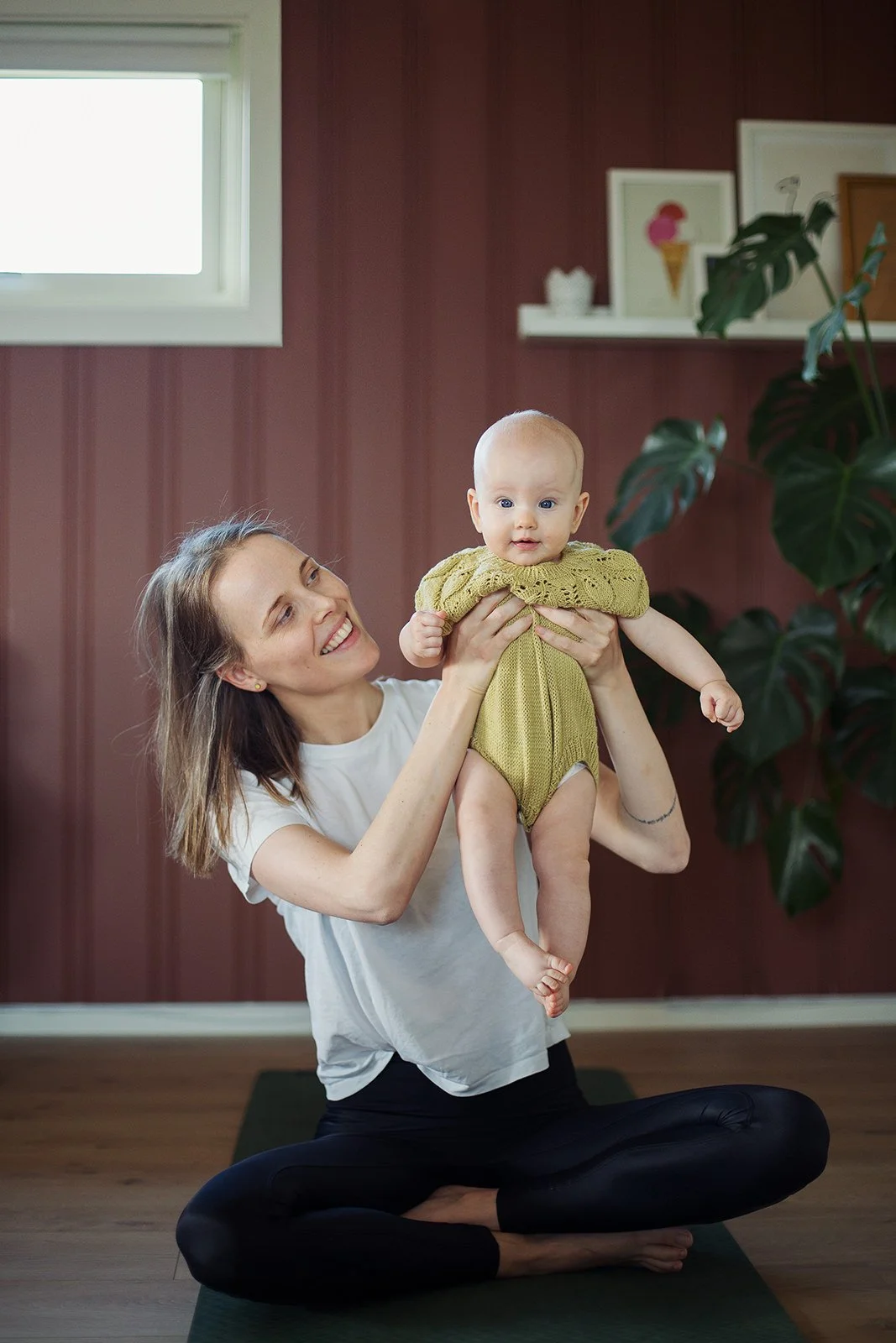 Mor og baby gjør yoga sammen. Sittende på gulvet løfter moren babyen opp.
