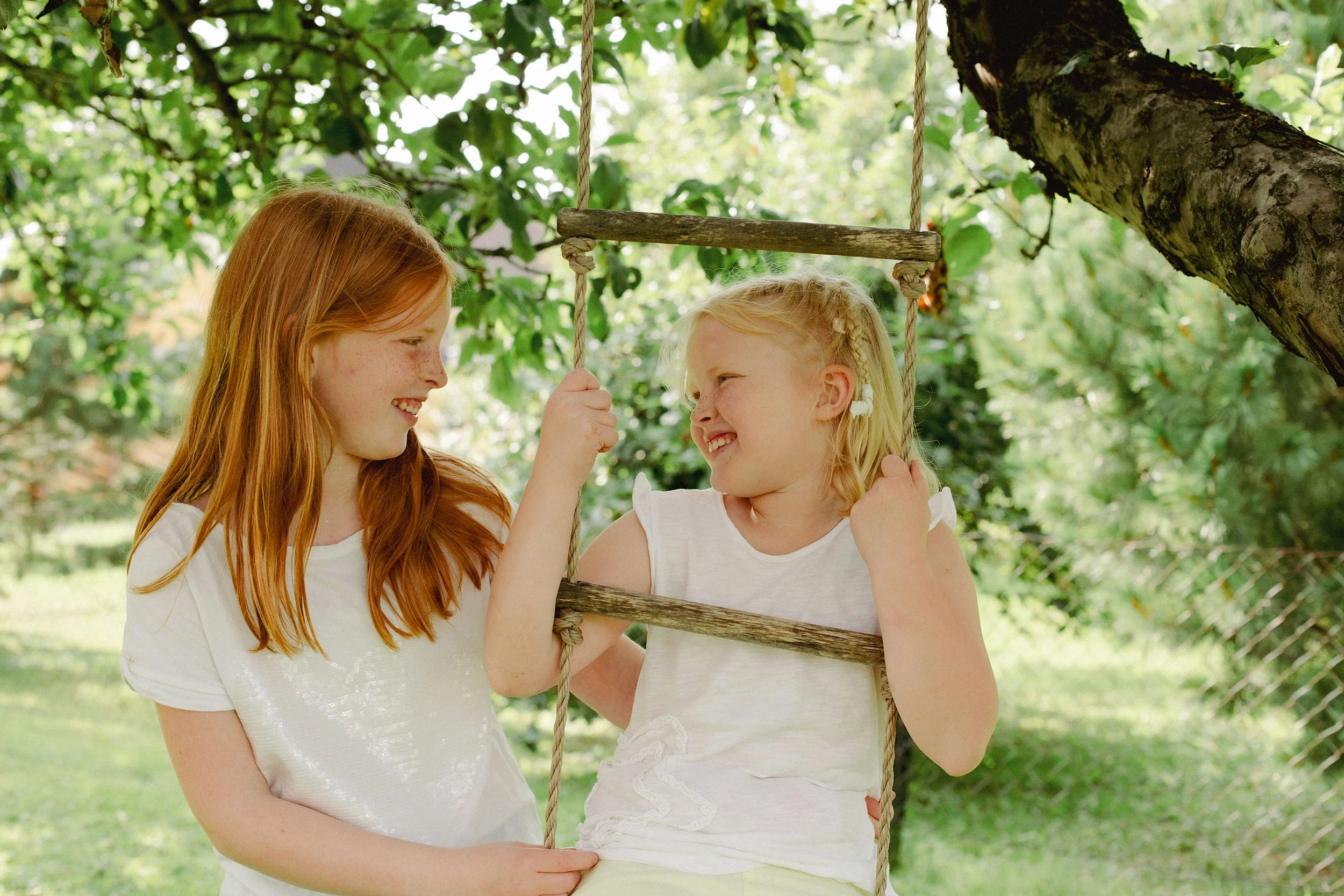 Familiefotografering utendørs i naturlig lys. Søsken, to søstre, smiler til hverandre.