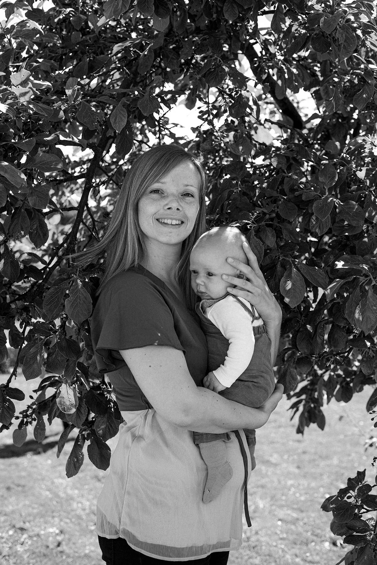 Mor holder baby i armene under skyggen av et tre på babyfotografering.