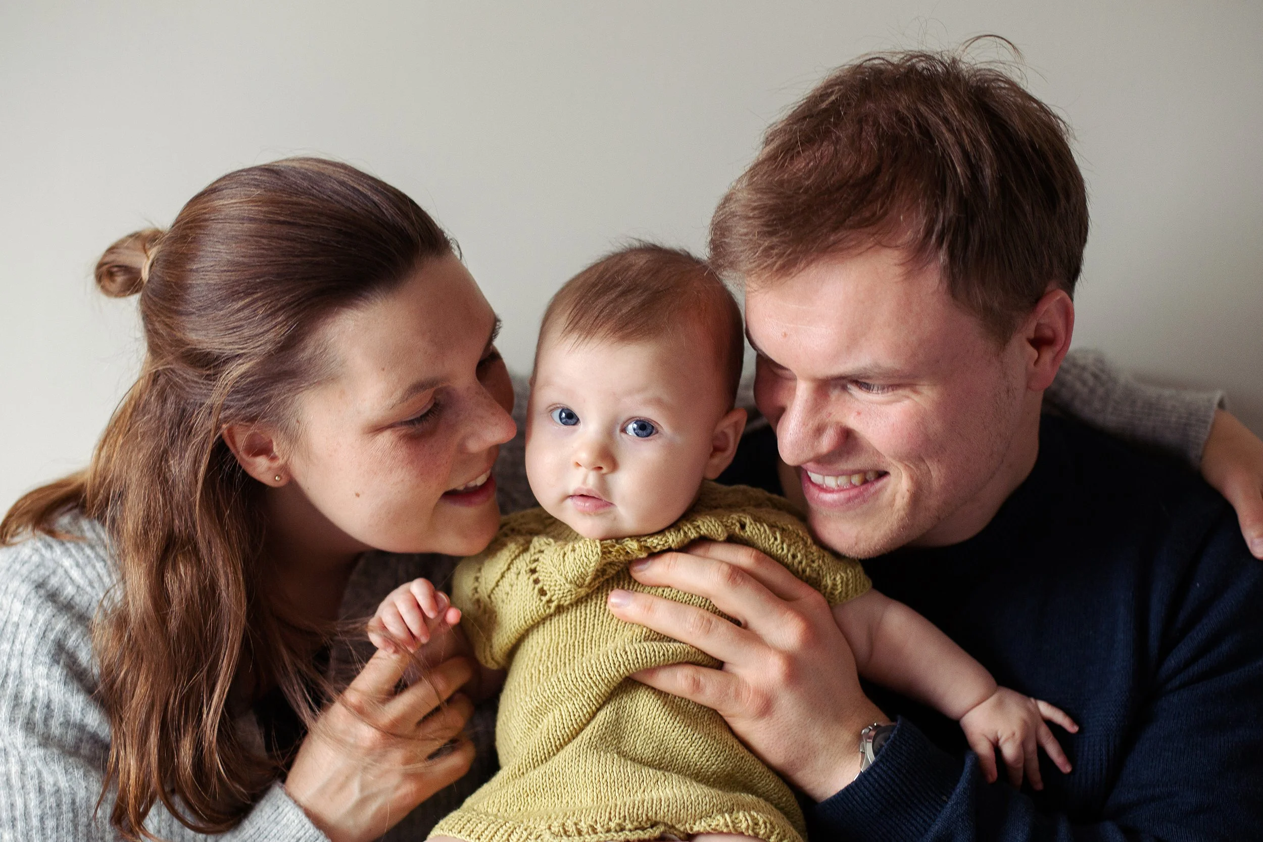 Familiefotografering innendørs i naturlig lys. Mor, far og baby på halvt år.