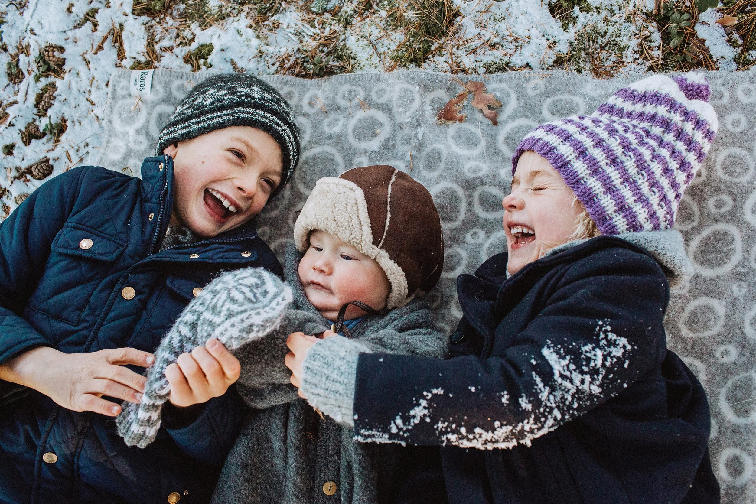 Tre barn ligger på et teppe i snøen og ler