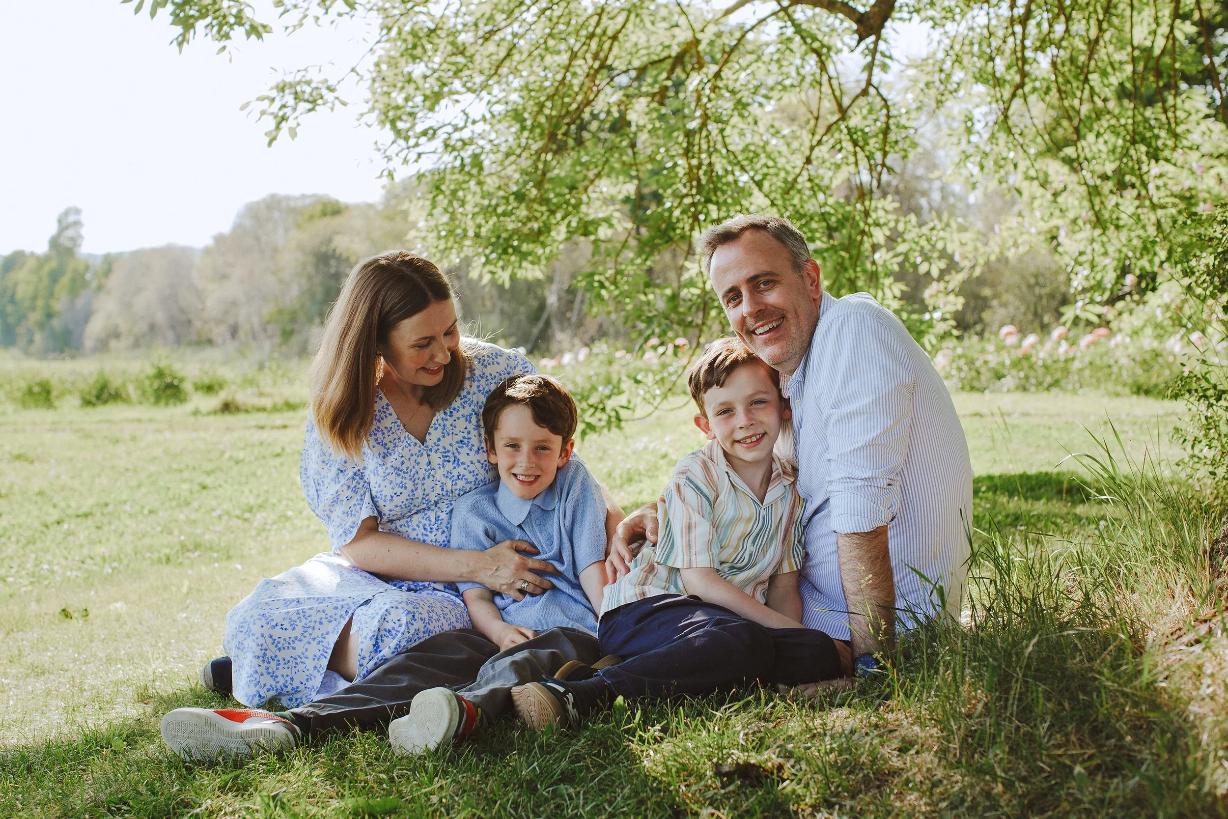 Familiefotografering av familie på fire som sitter under et tre på en eng på sommeren.