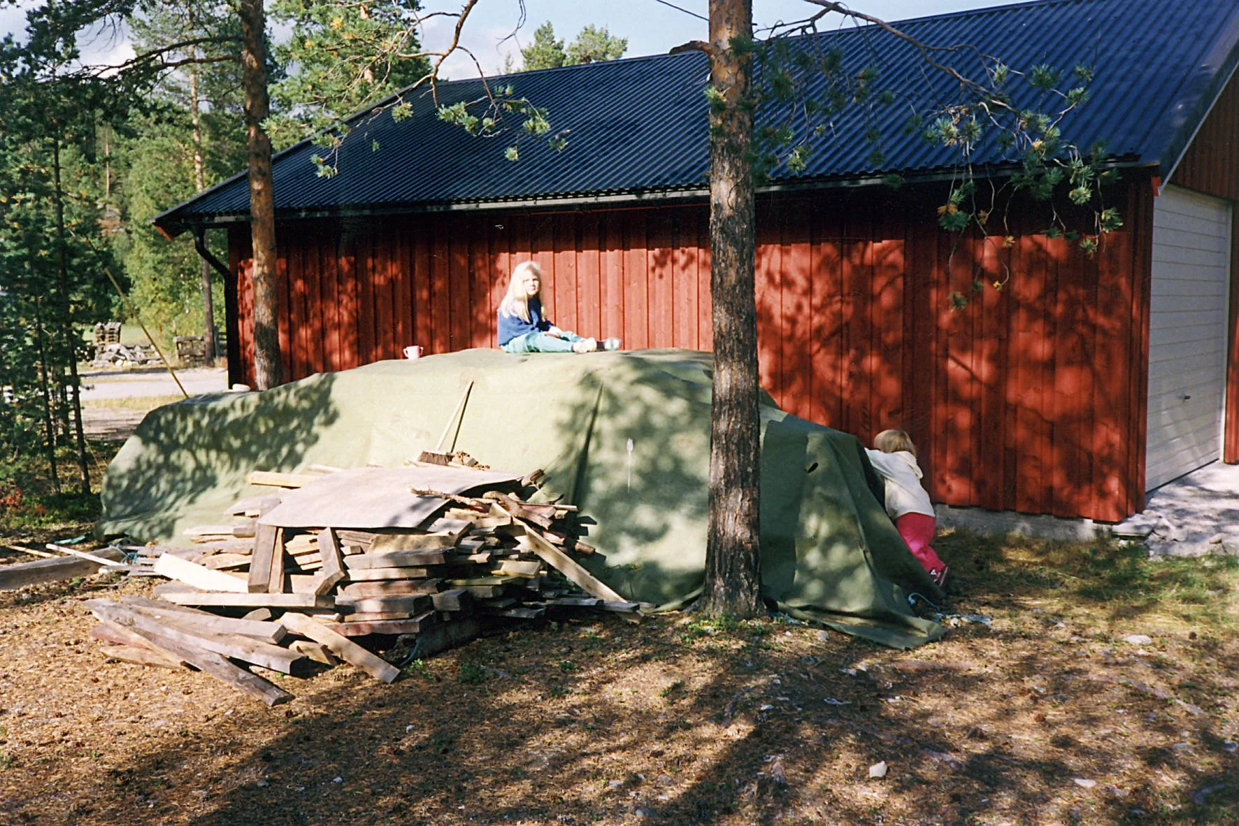 Digitalisert bilde av to barn som leker på en vedhaug dekket av presenning ved siden av en rød garasje.