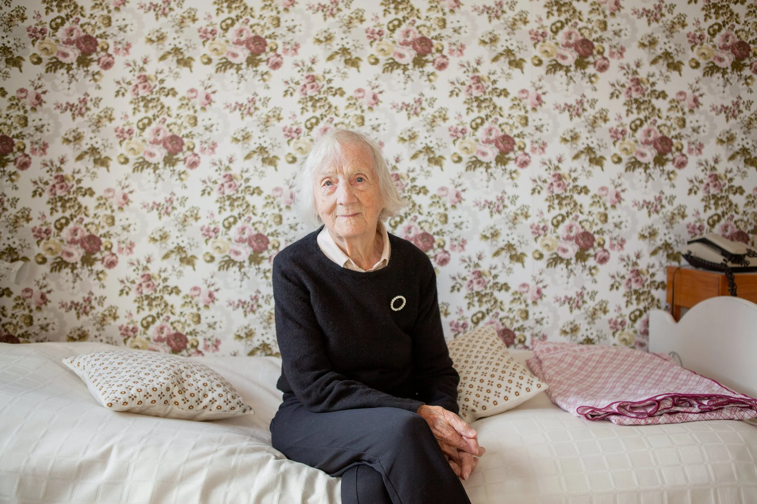 Portrettfotografering. Eldre kvinne sitter på sengen med blomstrete tapet på veggen bak.
