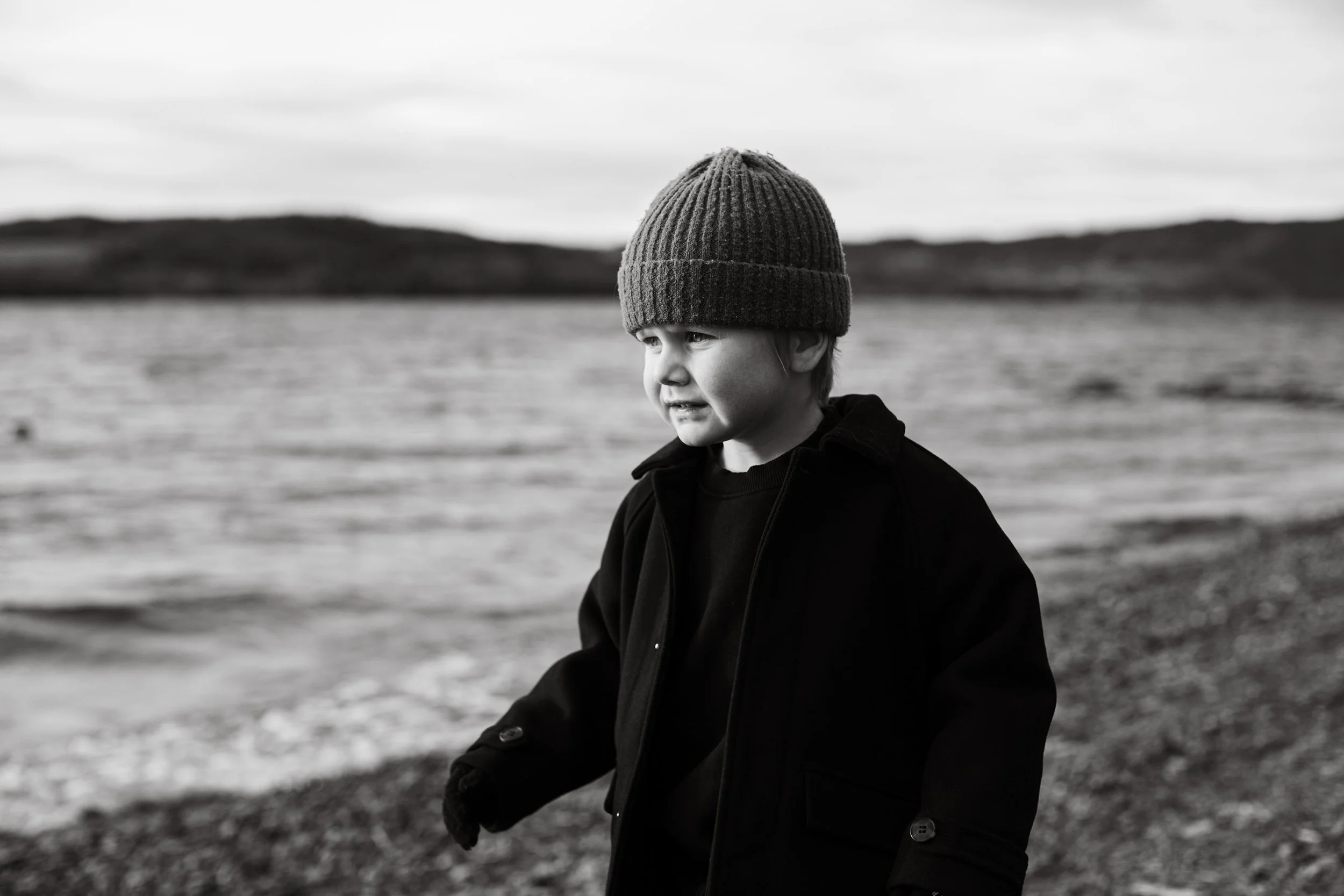 Barnefotografering på vinter. Gutt med lue står ved vannkanten på stranda.