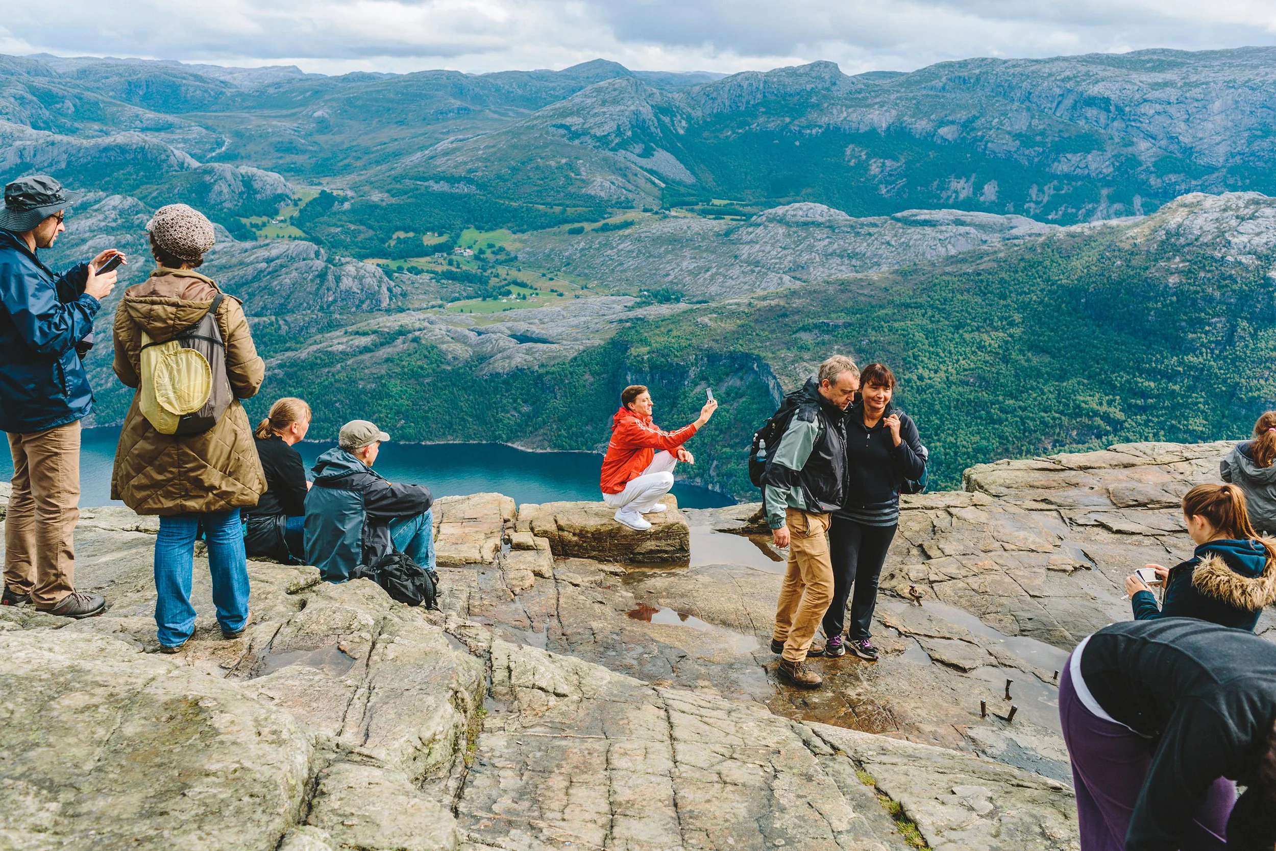 Mann tar selvportrett på huk på toppen av Prekestolen med mektig fjellandskap i bakgrunnen.