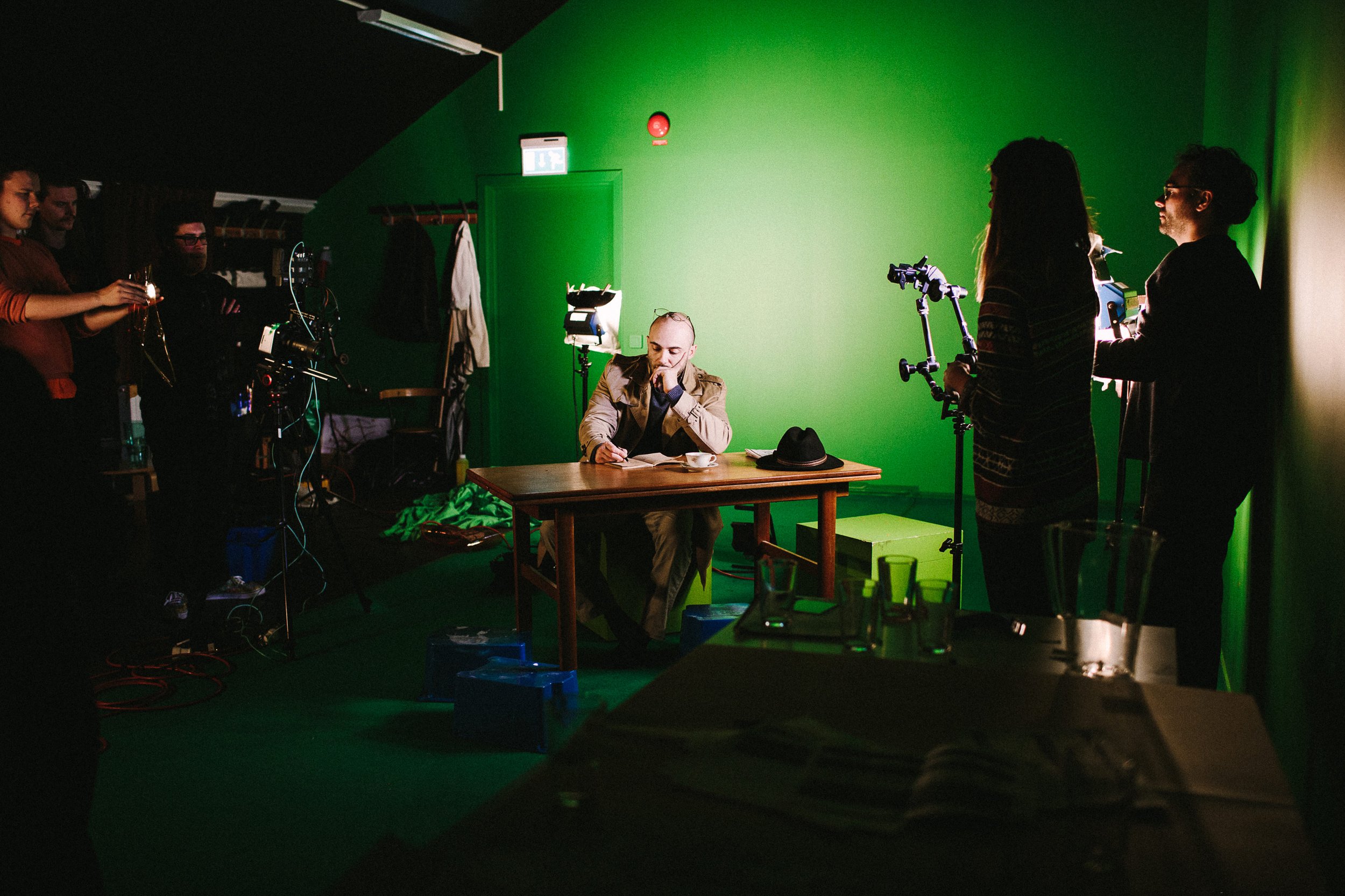 Skuespiller sittende ved et bord i et green screen rom under filminnspilling.