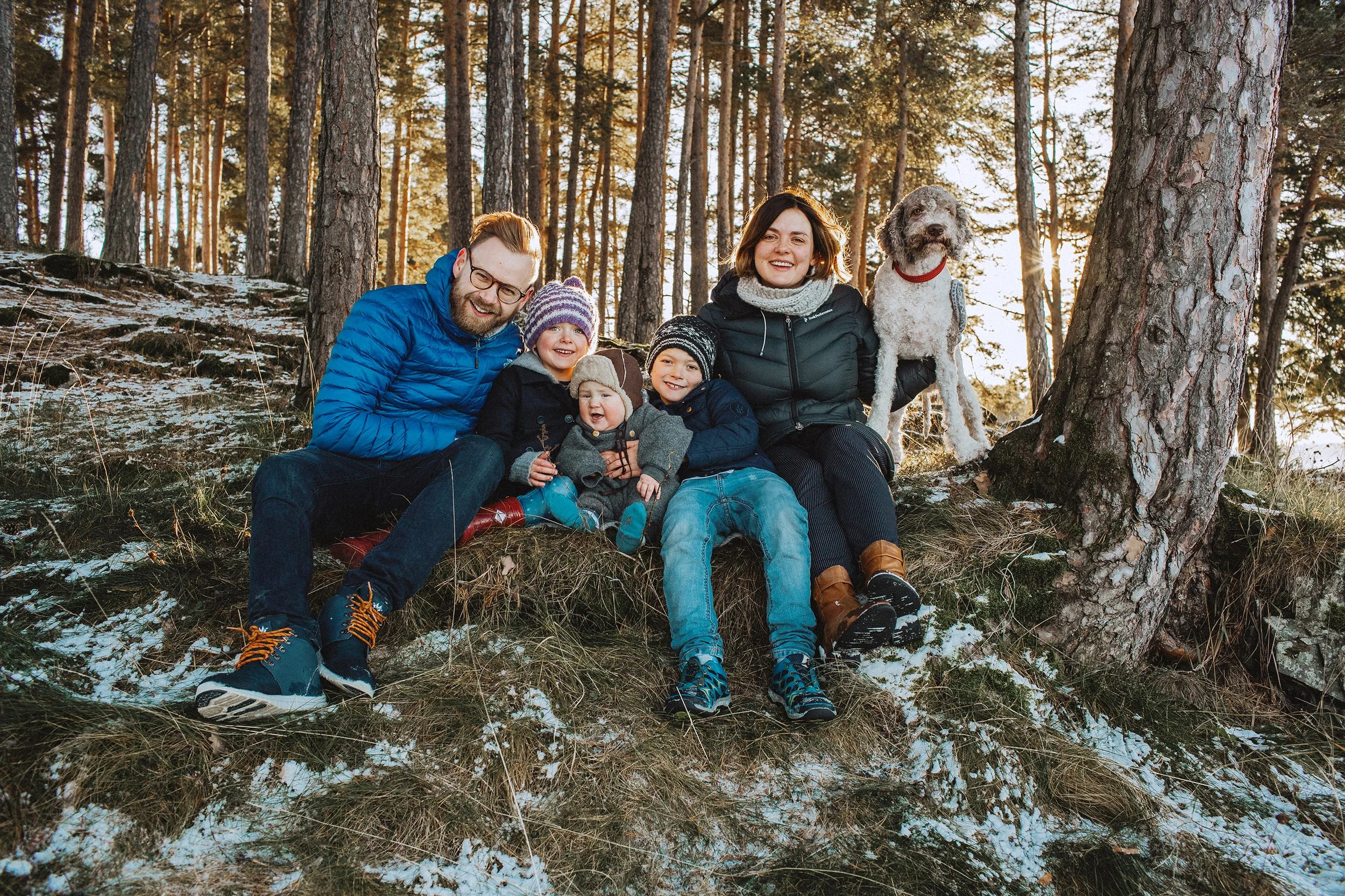 Familiefotografering utendørs på vinteren. Familie på fem og en hund sitter sammen på bakken i skogen.