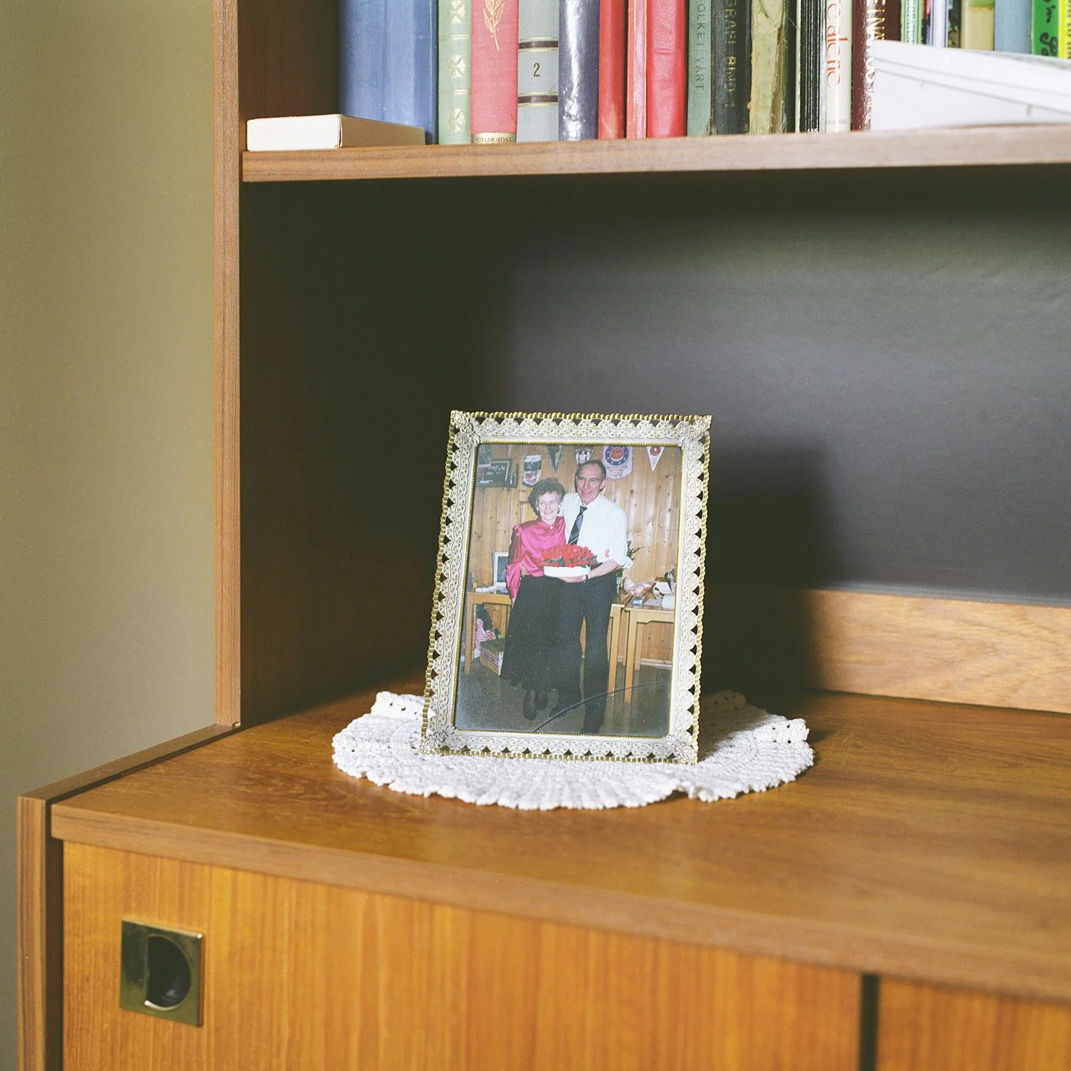 Utsnitt av interiør. Innrammet bilde av mann og kvinne som holder rundt hverandre. Rammen står på en heklet duk på en hylle.