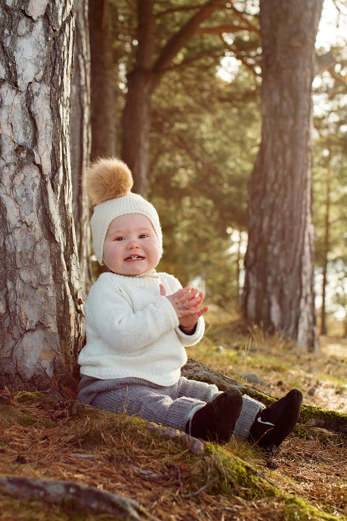 Barnefotografering om høsten. Barn sitter på bakken inntil et tre og smiler og klapper med hendene.