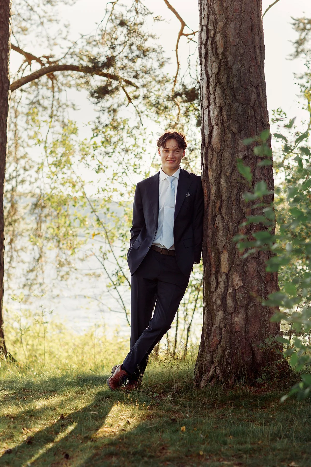 Konfirmantfotografering. Konfirmant i dress som avslappet lener skulderen inn mot et tre