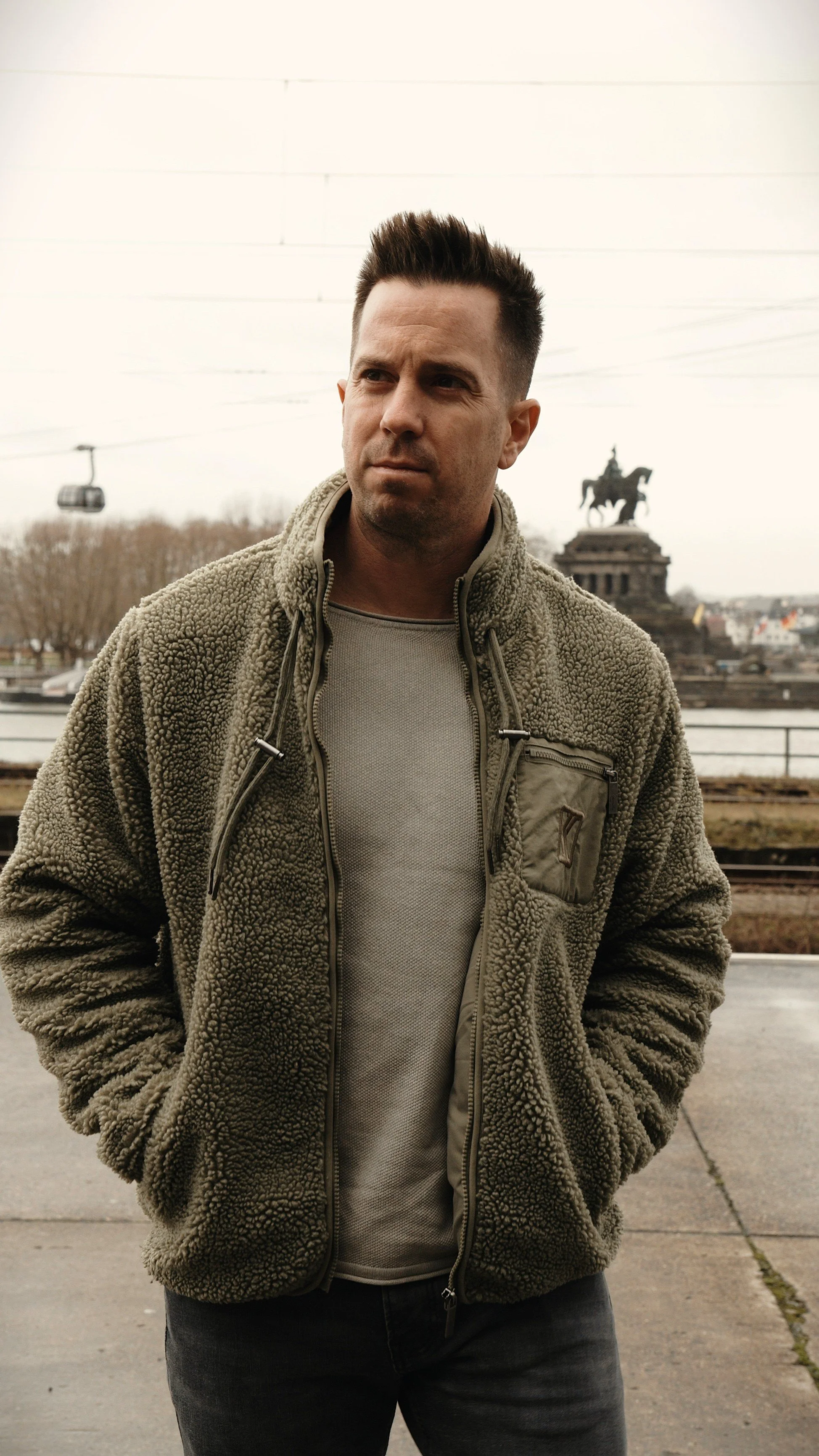 Junger Mann mit kurzen dunklen Haaren, trägt eine grüne, fleeceartige Jacke und graue Kleidung, steht vor einer städtischen Kulisse mit einer Statue auf einem Pferd im Hintergrund.