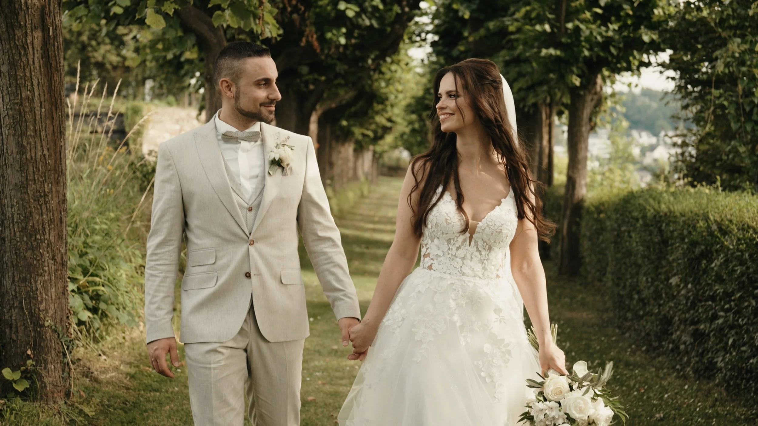 Ein Brautpaar, das Hand in Hand im Park spaziert, die Frau trägt ein weißes Hochzeitskleid und hält einen Blumenstrauß, der Mann trägt einen beigen Anzug mit Fliege.