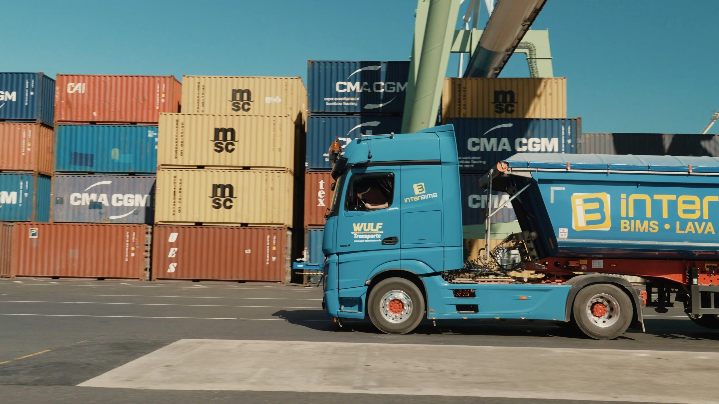 Ein blauer Lastwagen fährt an Container am Hafen vorbei, die Container sind in verschiedenen Farben und Marken wie CMA CGM, MSC und CAI gestapelt.