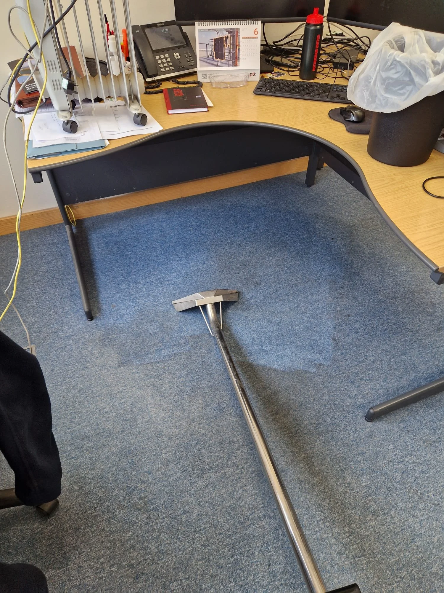 Office desk with a standing desk converter on the floor, a desktop monitor, keyboard, mouse, desk organizer, books, phone, calendar, water bottle, and garbage bin.