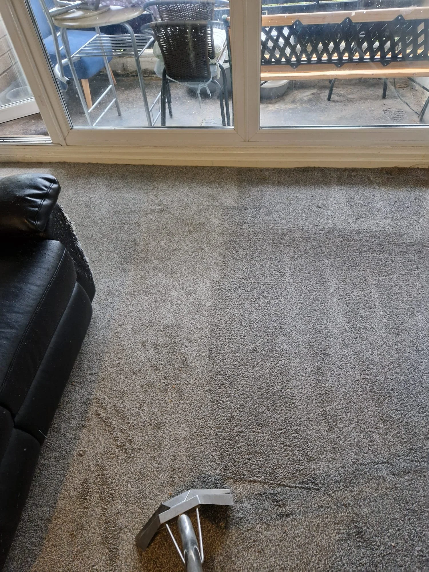 Living room with a large sliding glass door, outdoor patio furniture and a beige carpet. A black leather sofa is partially visible on the left, and a deodorizer spray bottle is on the floor.