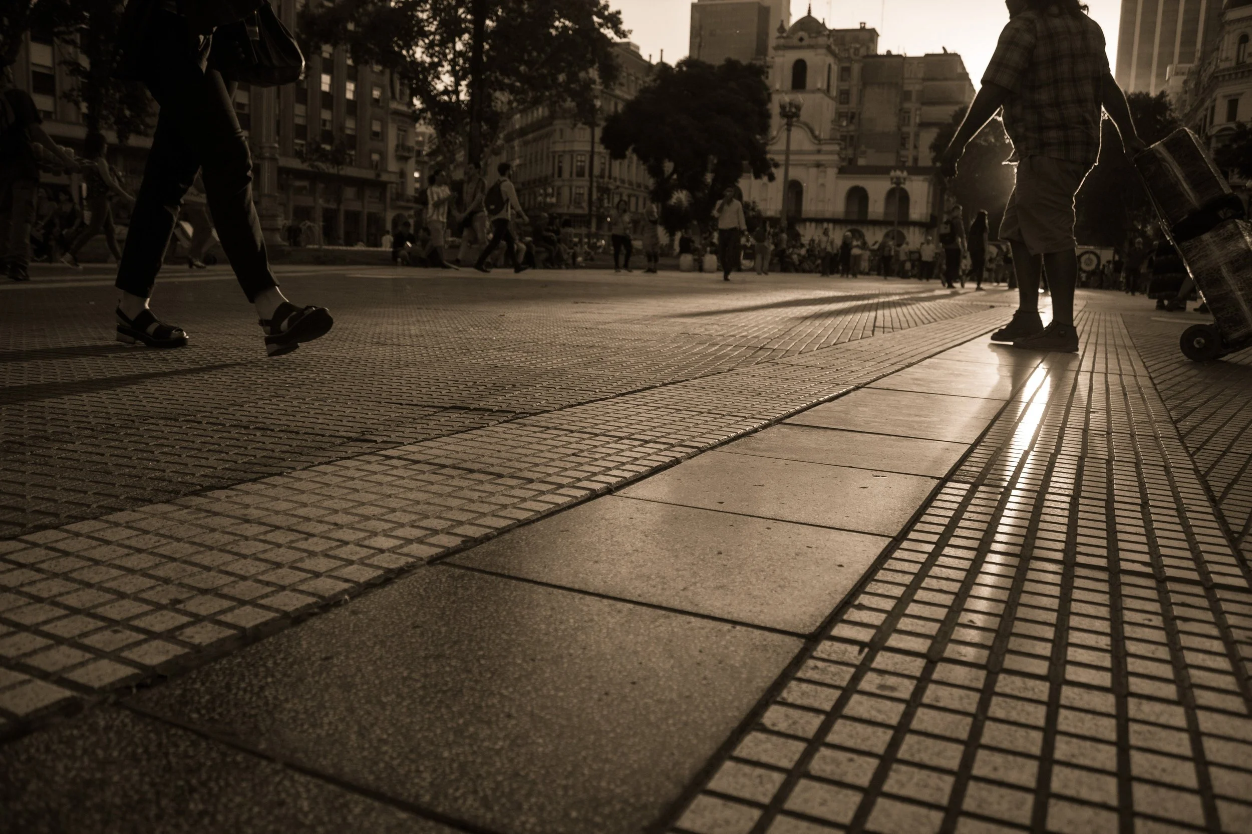 Silhouettes of people walking on a city street during sunset with historic buildings in the background.