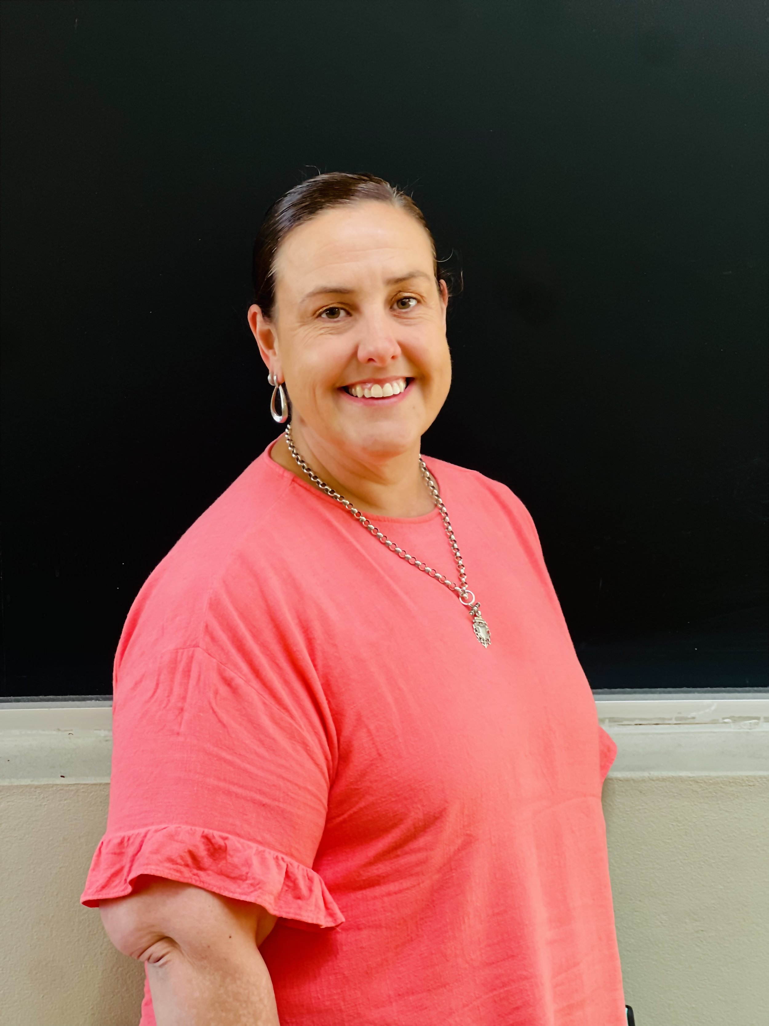 A smiling woman with short dark hair, wearing a red shirt with ruffled sleeves, a silver necklace with a pendant, and earrings, standing in front of a blackboard in a classroom.