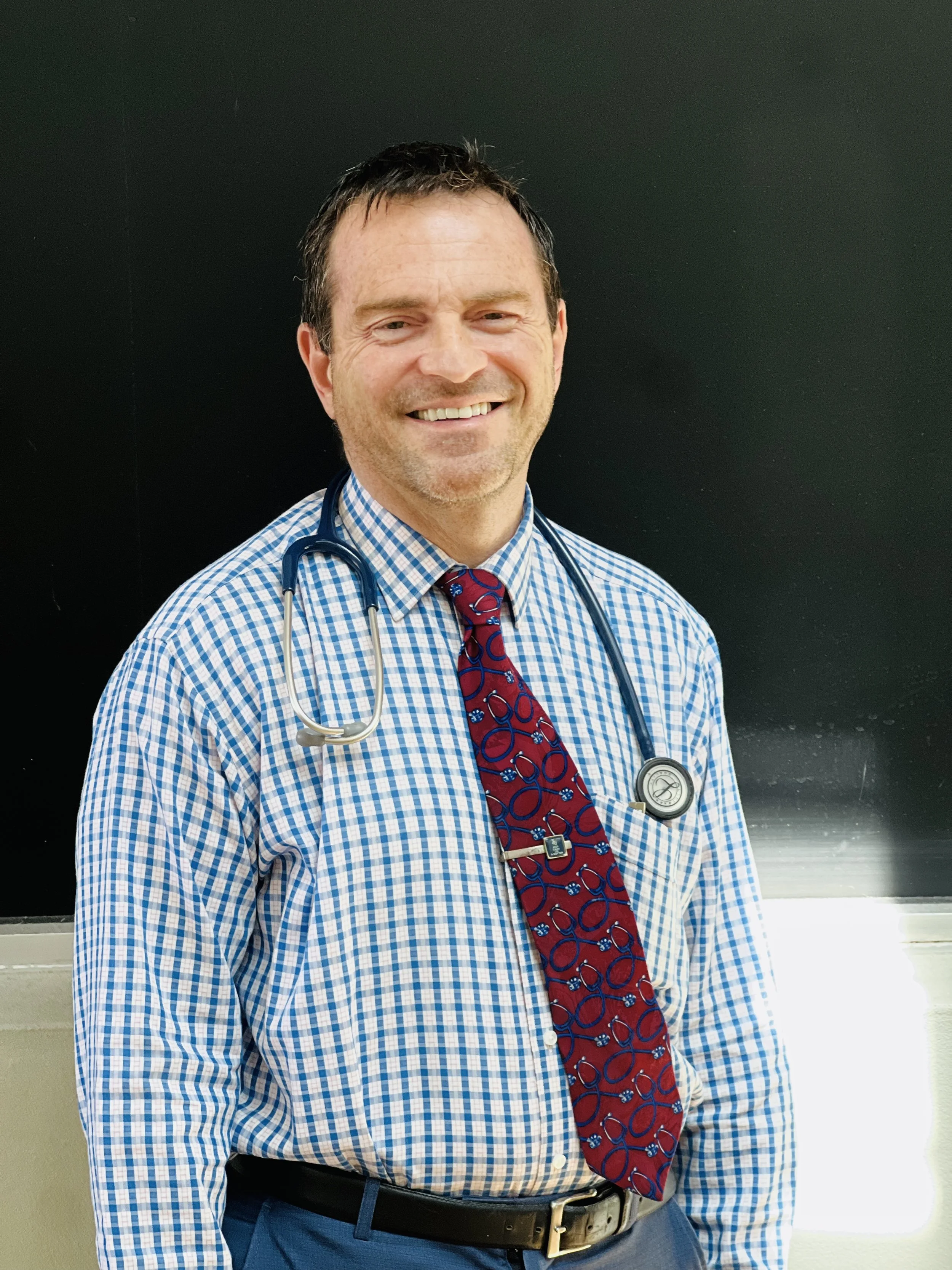 A smiling male doctor with brown hair, wearing a blue and white checkered shirt, a red patterned tie, and a stethoscope around his neck, standing in front of a black background.
