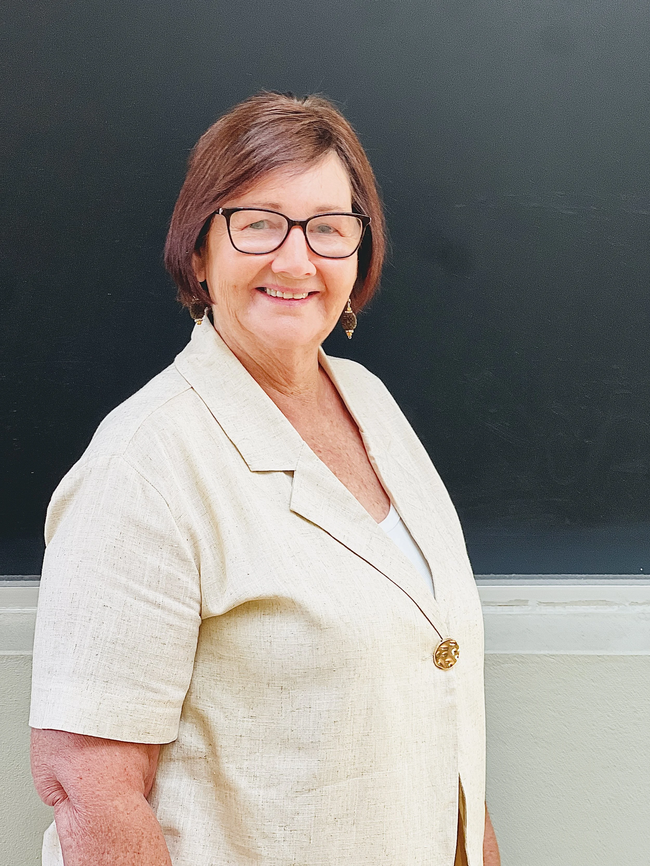 A smiling woman with glasses and short brown hair standing in front of a blackboard wearing a beige jacket and earrings.