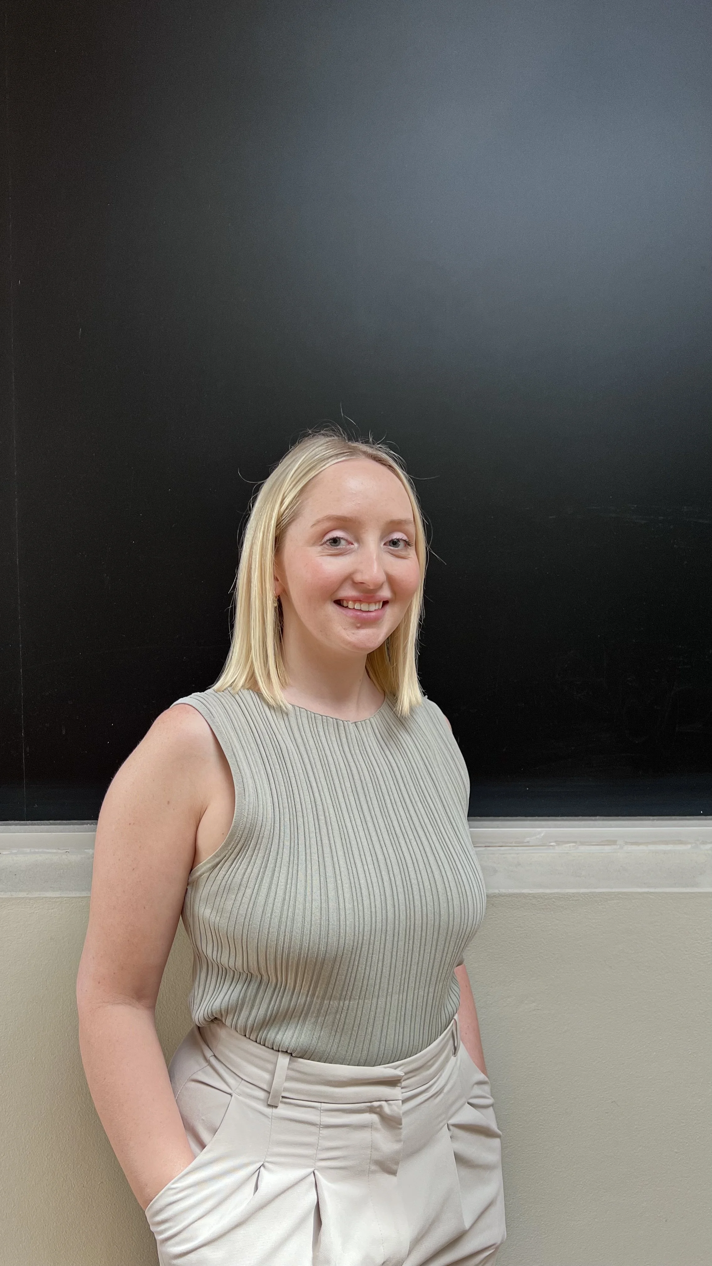 Young woman with blonde hair smiling, wearing a red sleeveless dress, in a parking lot with cars in the background.
