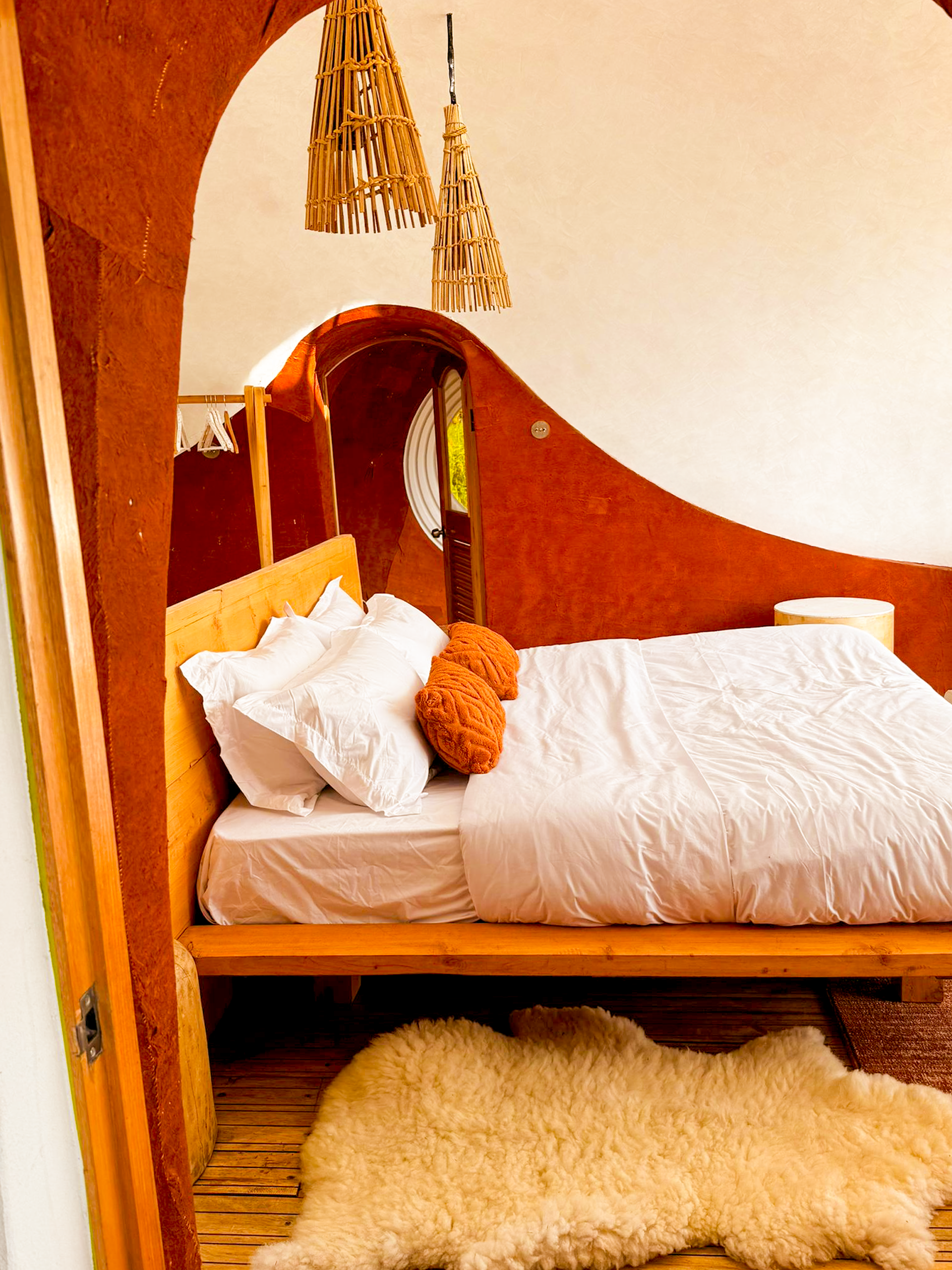 Cozy bedroom with wooden bed, white bedding, orange decorative pillows, sheepskin rug, wooden ceiling, bamboo pendant lights, and curved wooden wall with small window.