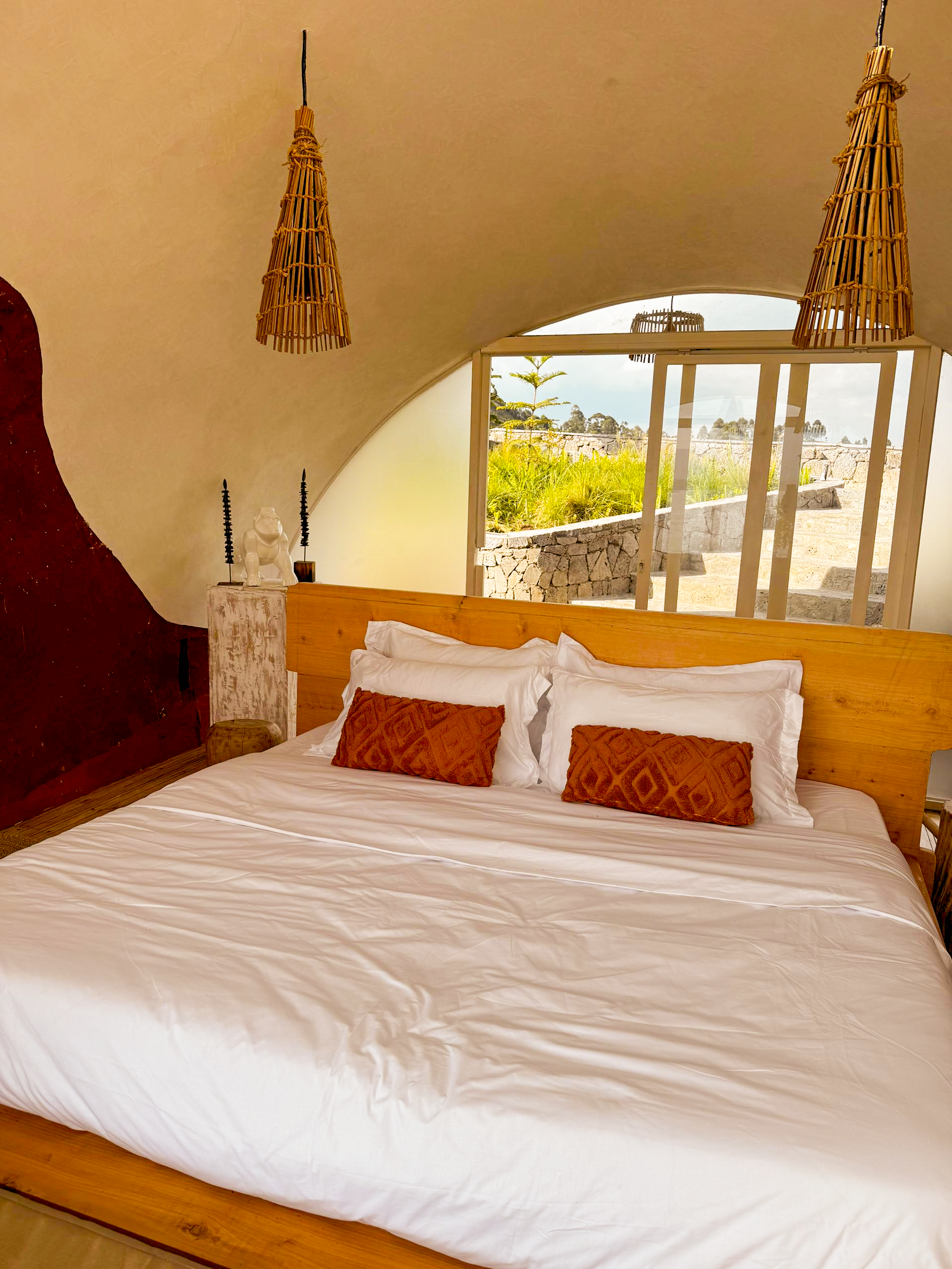 A cozy bedroom with a wooden bed frame, white bedding, and two orange decorative pillows. The room has a curved ceiling with two hanging wicker lamps and a large window with sliding glass doors showing an outdoor stone staircase, greenery, and a clou