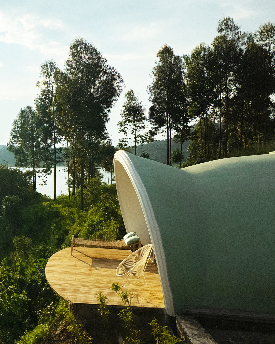 Modern house with curved roof on a hillside surrounded by trees, with outdoor seating including a lounge chair and a bench on a wooden deck.
