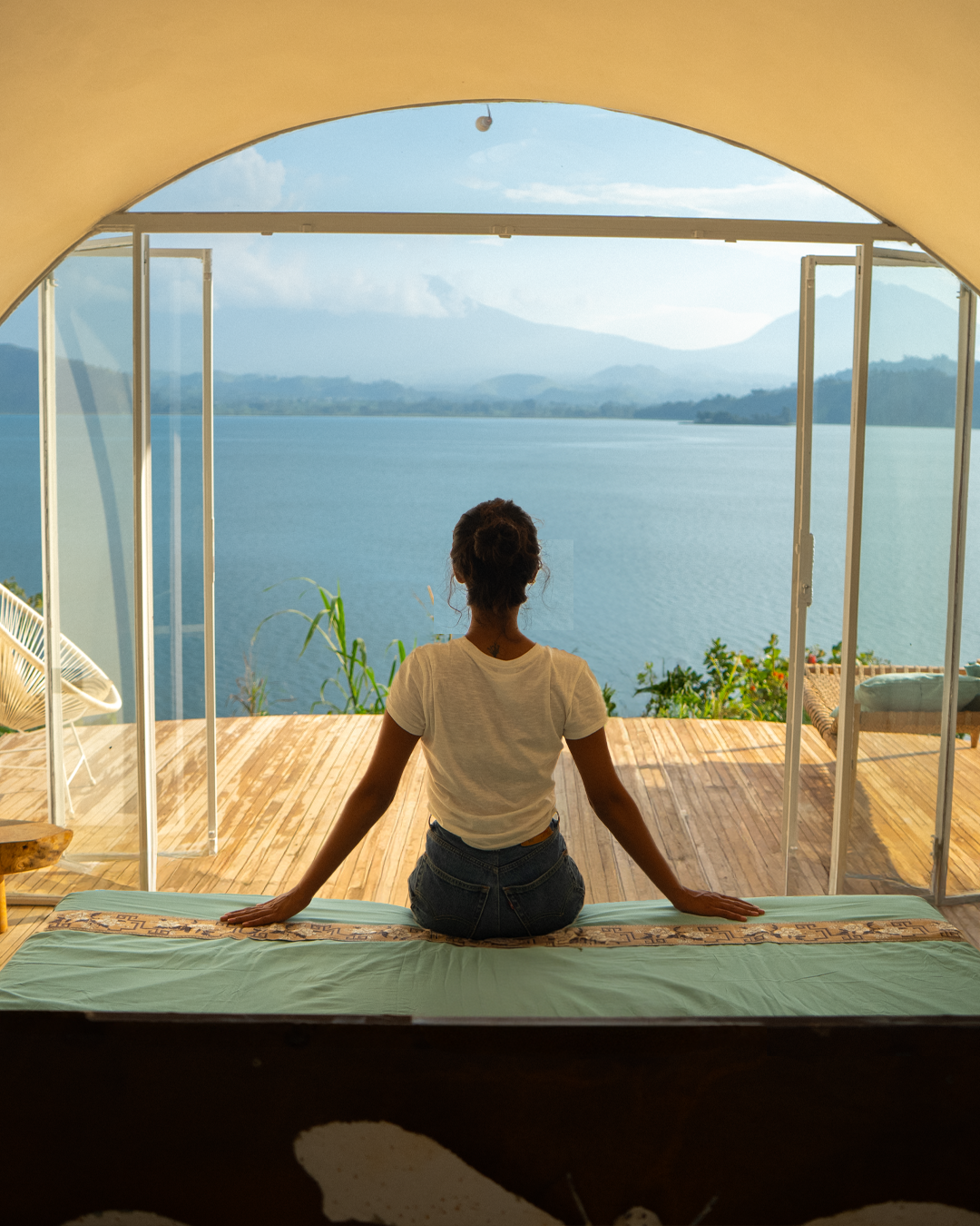 Woman sitting on a bed, looking out of open glass doors at a lake and mountains in the distance.